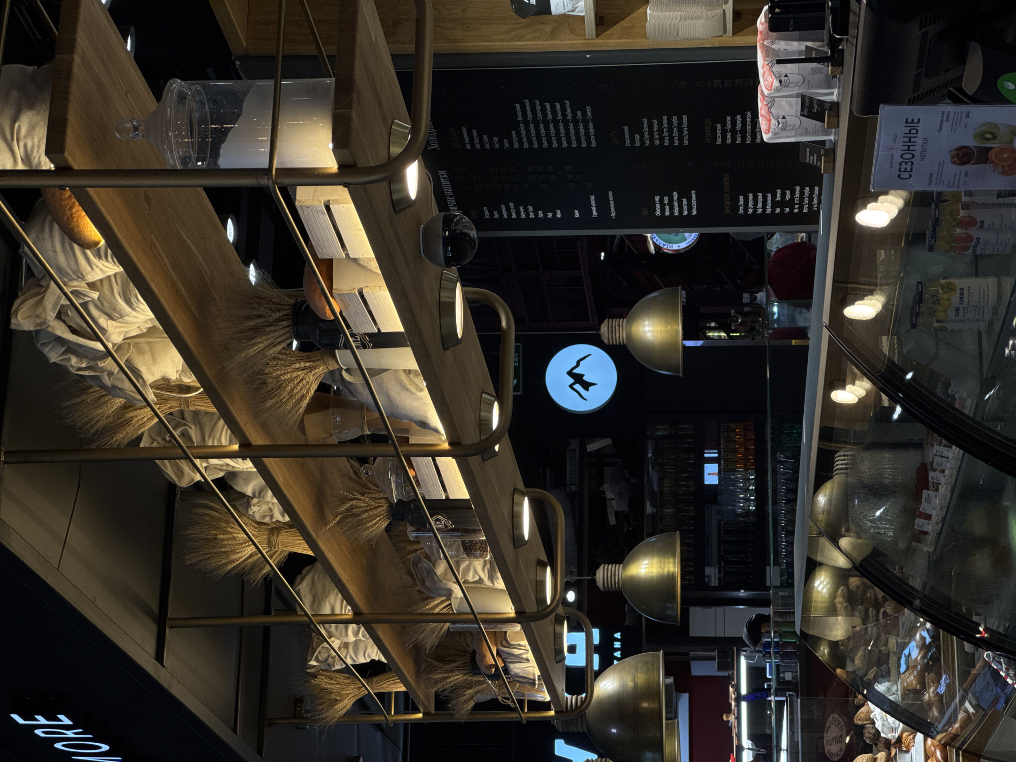 Here is a description of the image:
The image shows the interior of a coffee shop, possibly located in Moscow, Russia based on the menu. The camera is angled to capture both a display case with pastries and a portion of the menu board. The display case features seasonal drinks. A wooden shelf is decorated with wheat sheaves and covered with white cloth. Modern lighting fixtures illuminate the area. The shop's logo features a silhouette of a figure jumping over what appears to be a mountain. The OCR also indicates the name "ZUEGG", a possible reference to a food brand and a sign that indicates exit. The perspective of the image makes it seem like the viewer is looking upwards within the cafe, capturing architectural and decorative elements. The overall feel is contemporary with a touch of rustic charm.