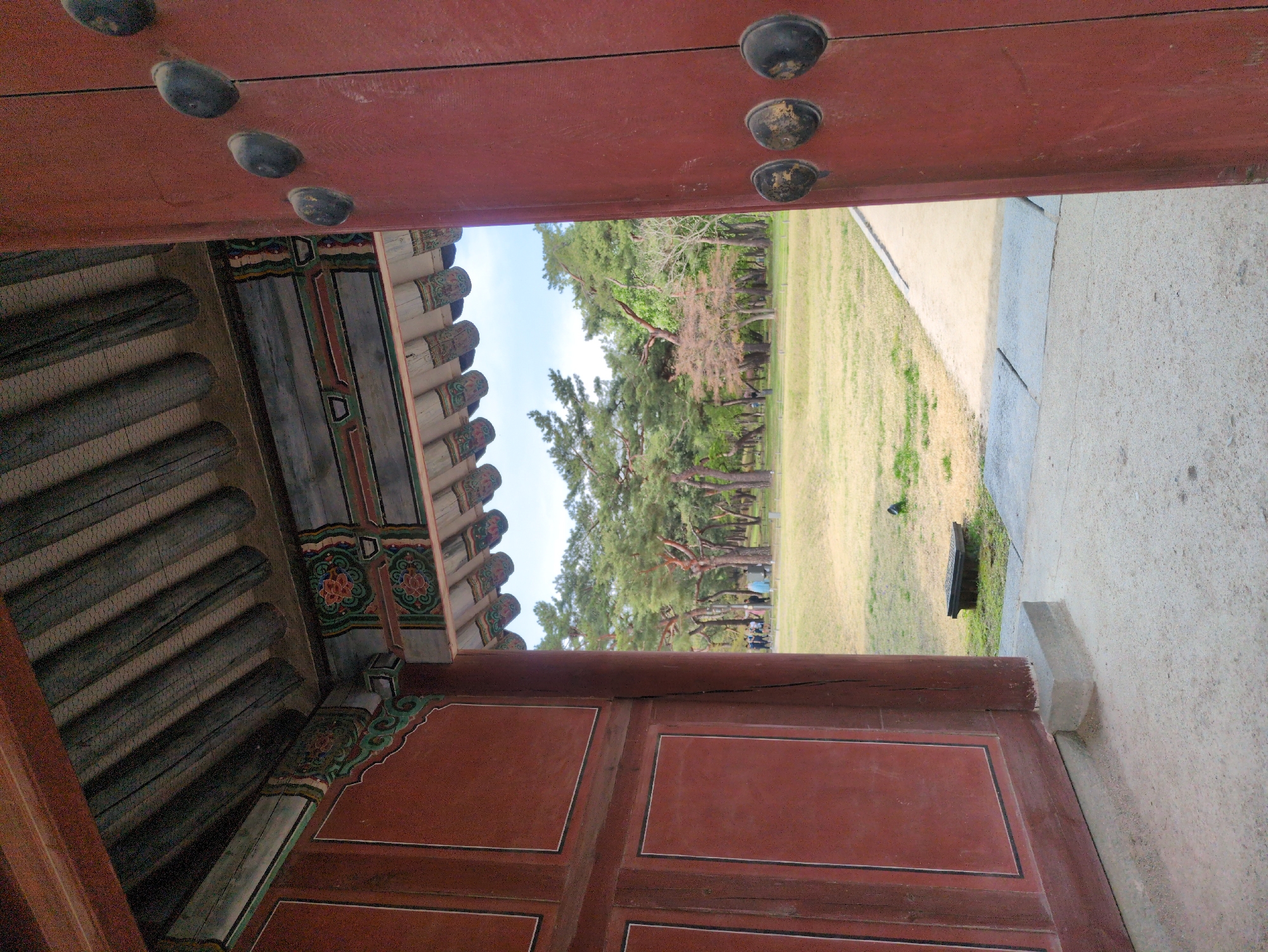 The image depicts a partial view through the open doors of a traditional Korean structure, possibly located in Seoul, South Korea, given the architectural style. The foreground shows the red-painted wooden door frame with decorative nails and panels. The interior of the structure is visible, showcasing the intricate design of the roof and support beams, adorned with traditional Korean patterns and colors. Through the opening, a view of a grassy area with trees is seen, suggesting an outdoor garden or park setting. People are visible in the distance. A concrete pathway is adjacent to the grass.