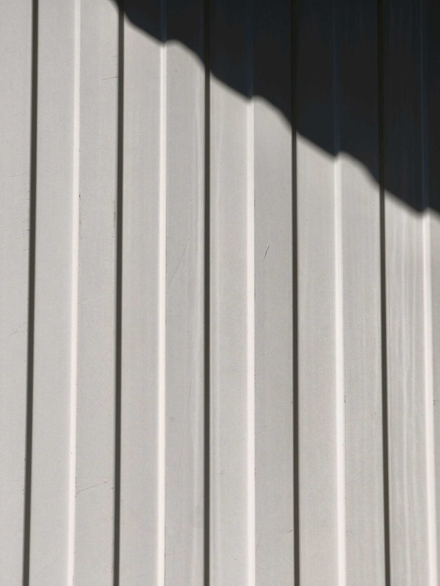 The image is a close-up, abstract view of a light-colored, corrugated metal surface, likely an exterior wall. The surface exhibits vertical ridges and valleys, creating a pattern of light and shadow. A diagonal shadow, cast by an unseen object, cuts across the upper right portion of the frame, highlighting the texture and dimensionality of the metal. The lighting suggests it is daytime. There are no people or discernible objects in the frame beyond the wall itself. The image is devoid of any specific identifiers that would place it in Karachayevsk, Russia, or any other specific city.