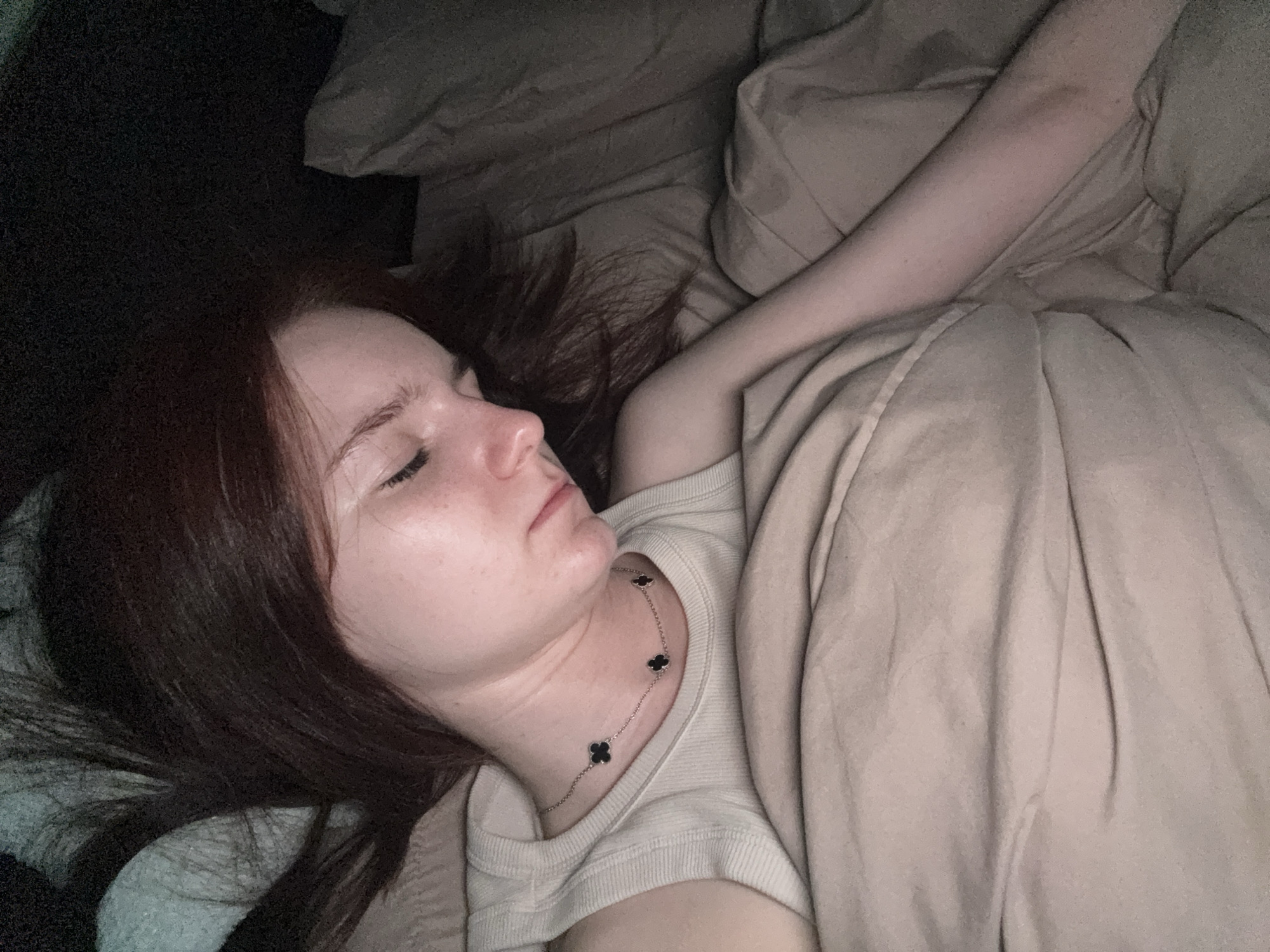 A person is sleeping in a bed. The person has long, dark hair and is wearing a light-colored top and a necklace with black clover-shaped pendants. The person is lying under a light beige blanket. The location cannot be determined from the image.
