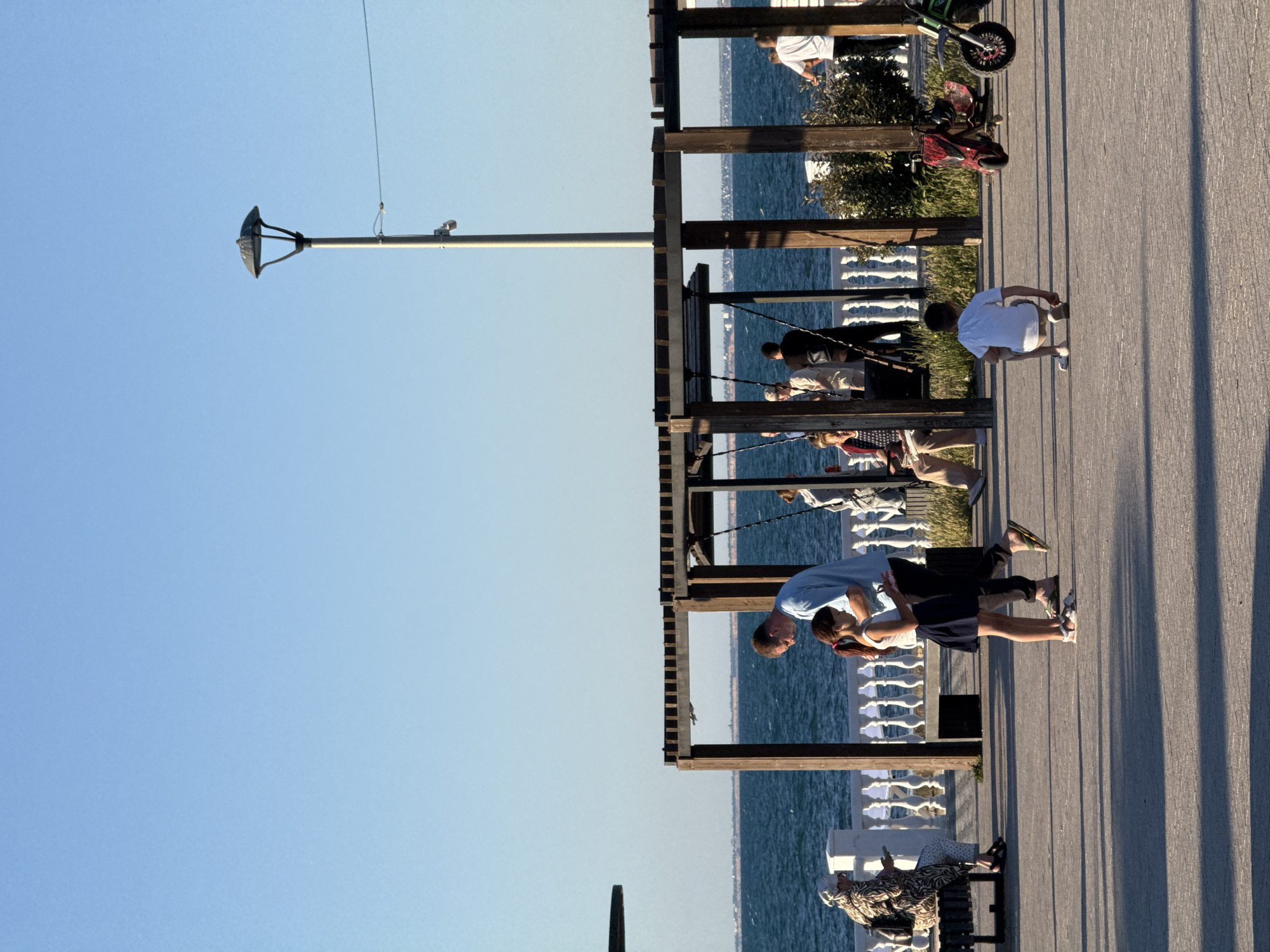 A wide shot captures a sunlit waterfront promenade with a clear blue sky and calm sea. In the foreground, a boy in a white t-shirt and shorts crouches on the pavement. To his left, a man in a light blue shirt and dark pants is talking to a young girl in a dark skirt and white top. Behind them, a woman wearing a patterned dress and headscarf sits on a bench. Further back, a wooden canopy structure with hanging swings is visible, occupied by several people. A lamppost stands tall to the right of the swings. On the far right, two electric scooters, one red and one green, are parked. The promenade is paved, and a white ornate railing separates it from the sea. Shadows from the canopy and people are cast on the ground, indicating it is daytime.