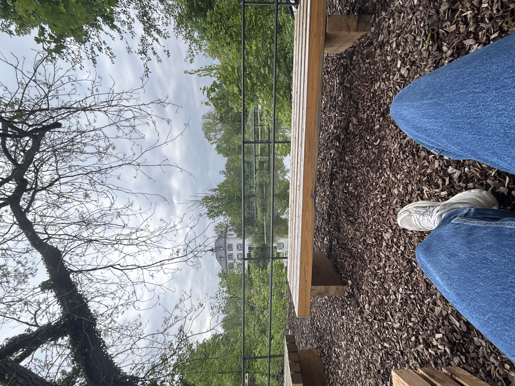A first-person perspective shot of a person sitting on a wooden bench in a park setting. The person is wearing blue jeans and a white sneaker. The ground is covered in brown mulch. A black metal railing separates the bench from a body of water, likely a river or lake. Trees and foliage are visible in the background. A white building is reflected in the water. The sky is overcast. The location appears to be in a natural setting within the city.