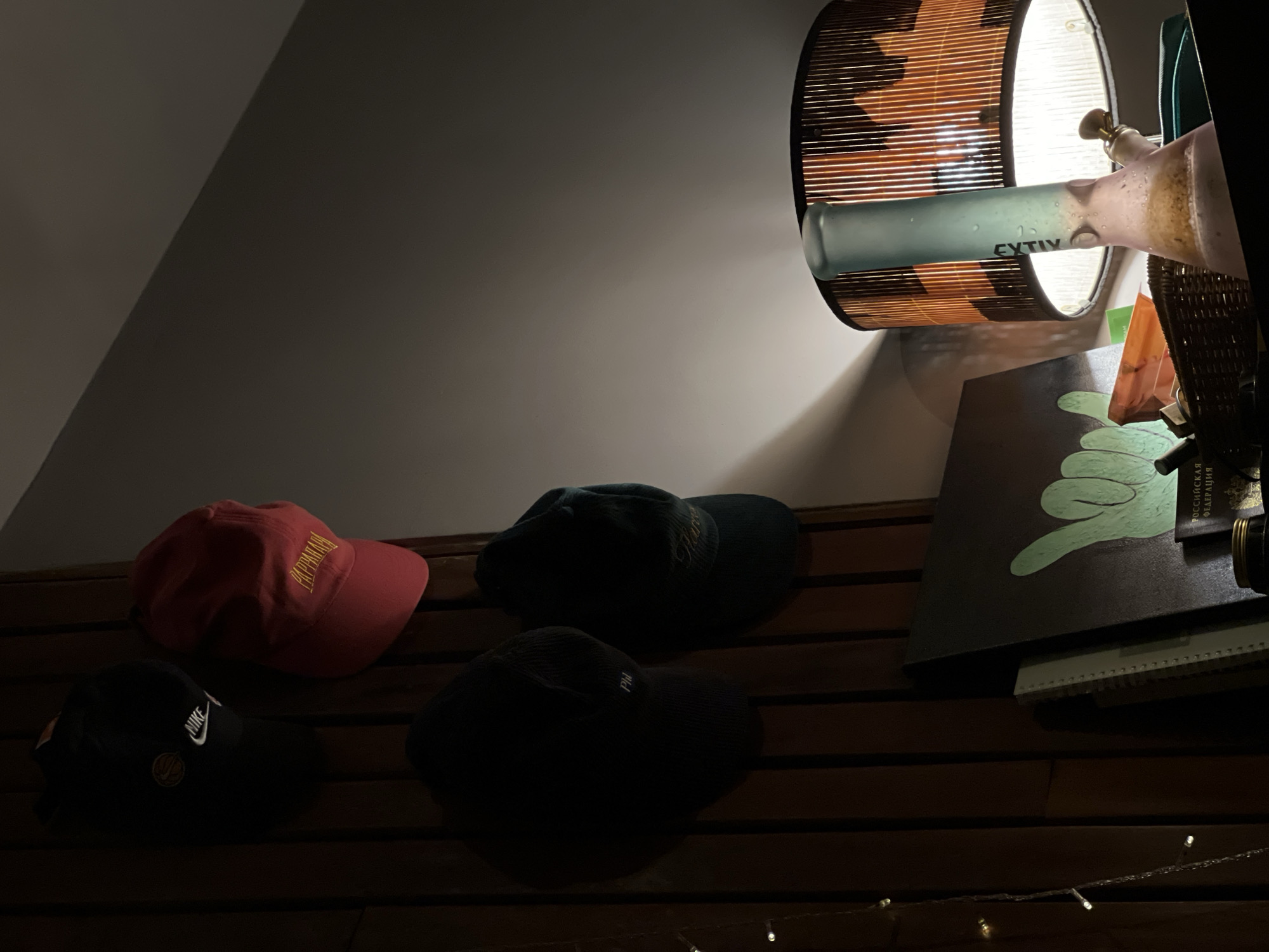 The image displays a dimly lit indoor scene, likely at night, focusing on a collection of personal items. A lamp with a cylindrical, slatted wooden shade, possibly made of bamboo, casts a warm light downwards and onto an angled, light-colored wall.
Prominently positioned in front of the lamp is a frosted glass object resembling a bong or water pipe, with a greenish lower half and a pinkish upper half, along with a brass-colored fitting. The word "GYTIV" is visible on the green portion of this object. To the immediate right of the lamp, hints of a woven basket and other dark items are visible.
Below the lamp and glass object, several baseball caps are arranged on a dark, horizontally slatted wooden surface, possibly a shelf or headboard. These include a red cap with faint yellow text that appears to say "PAPPALU", a black cap featuring the white Nike swoosh logo, and two other dark-colored caps, one of which shows visible initials "PM" in light embroidery.
Further to the right, a dark rectangular object with a stylized light green drawing of a hand in a "shaka" or "hang loose" gesture rests on top of what appear to be books or notebooks. Partially visible Russian text on this object reads "РОССИЙСКАЯ" (Russian) and "ФЕДЕРАЦИЯ" (Federation). Along the very bottom edge of the frame, a string of small, warm white fairy lights adds additional soft illumination to the scene. No people are visible, and the scene depicts a static arrangement of items within a personal space.