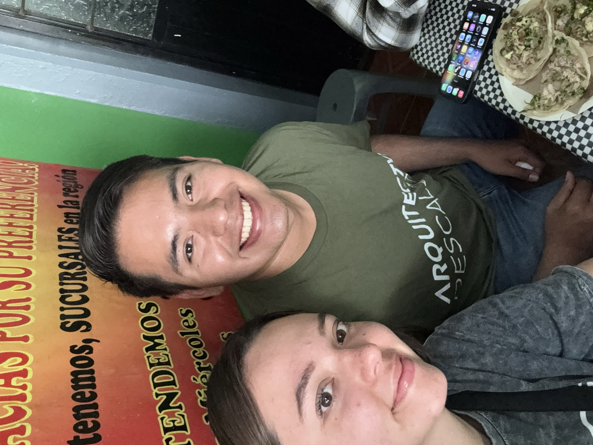 Here is a description of the image:
The image depicts a couple taking a selfie indoors, likely in a restaurant or establishment. To their side is a promotional sign with the words "SOMOS POR SU PREFERENCIA tenemos, SUCURSALES en la región TENDEMOS" visible in part. The location is suggested to be in Cuetzalan, Mexico.
On the table, next to the couple, there's a plate of what appears to be tacos, and a smartphone. The man is wearing an olive-green t-shirt with a white graphic or text on it. The woman is wearing a gray t-shirt. Both are smiling at the camera. The background includes what looks like a green wall and part of the ceiling. A checkerboard pattern table is near the top edge of the frame. A gray stool also visible.