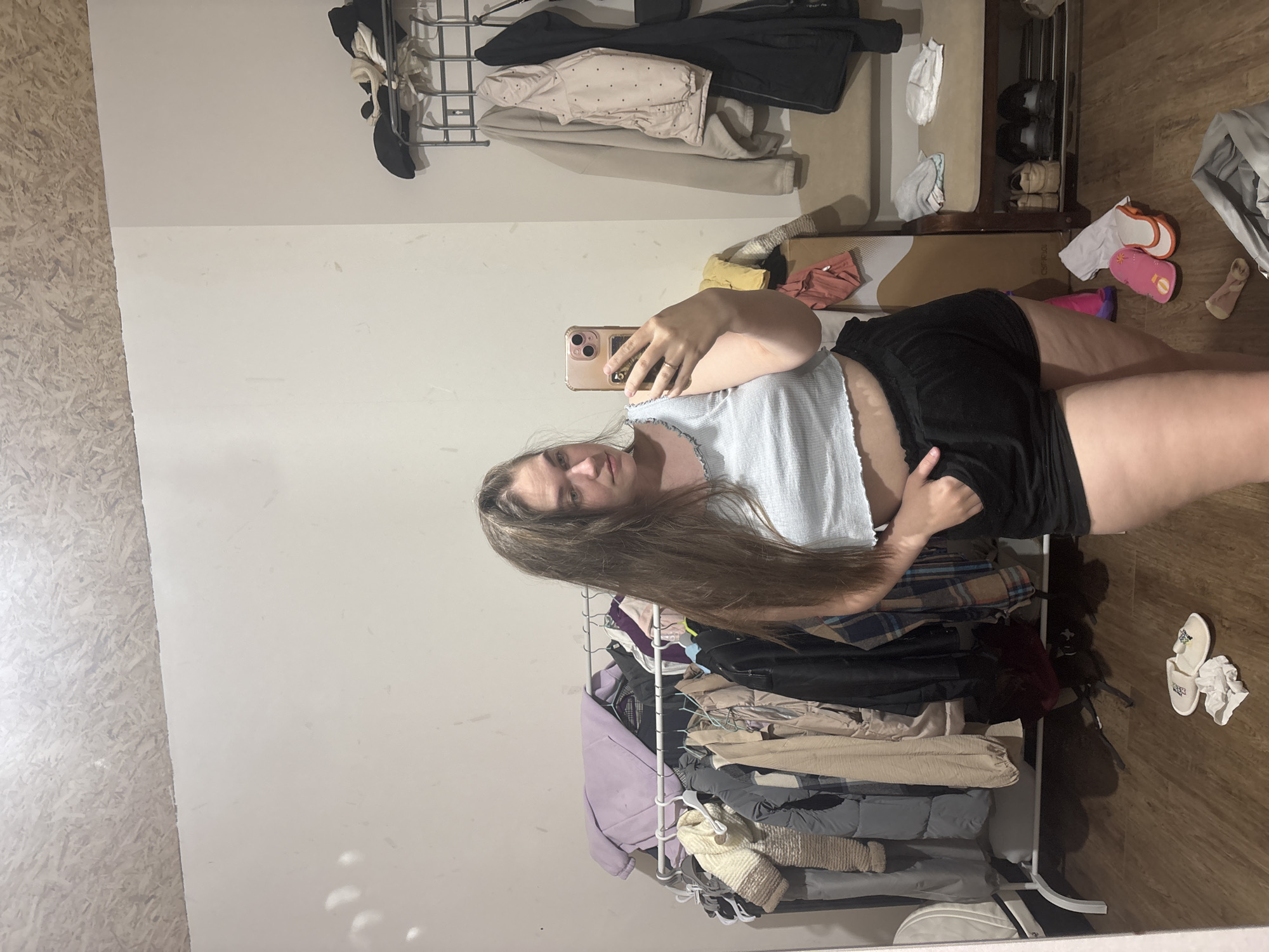 A young woman with long brown hair is taking a selfie in a room. She is wearing a light blue top and black shorts. Behind her is a rack of clothing, and various items are scattered on the floor. The setting appears to be a room with light-colored walls and wooden flooring. There is no information to suggest the image was taken in the city of Chaikovsky, Russia.