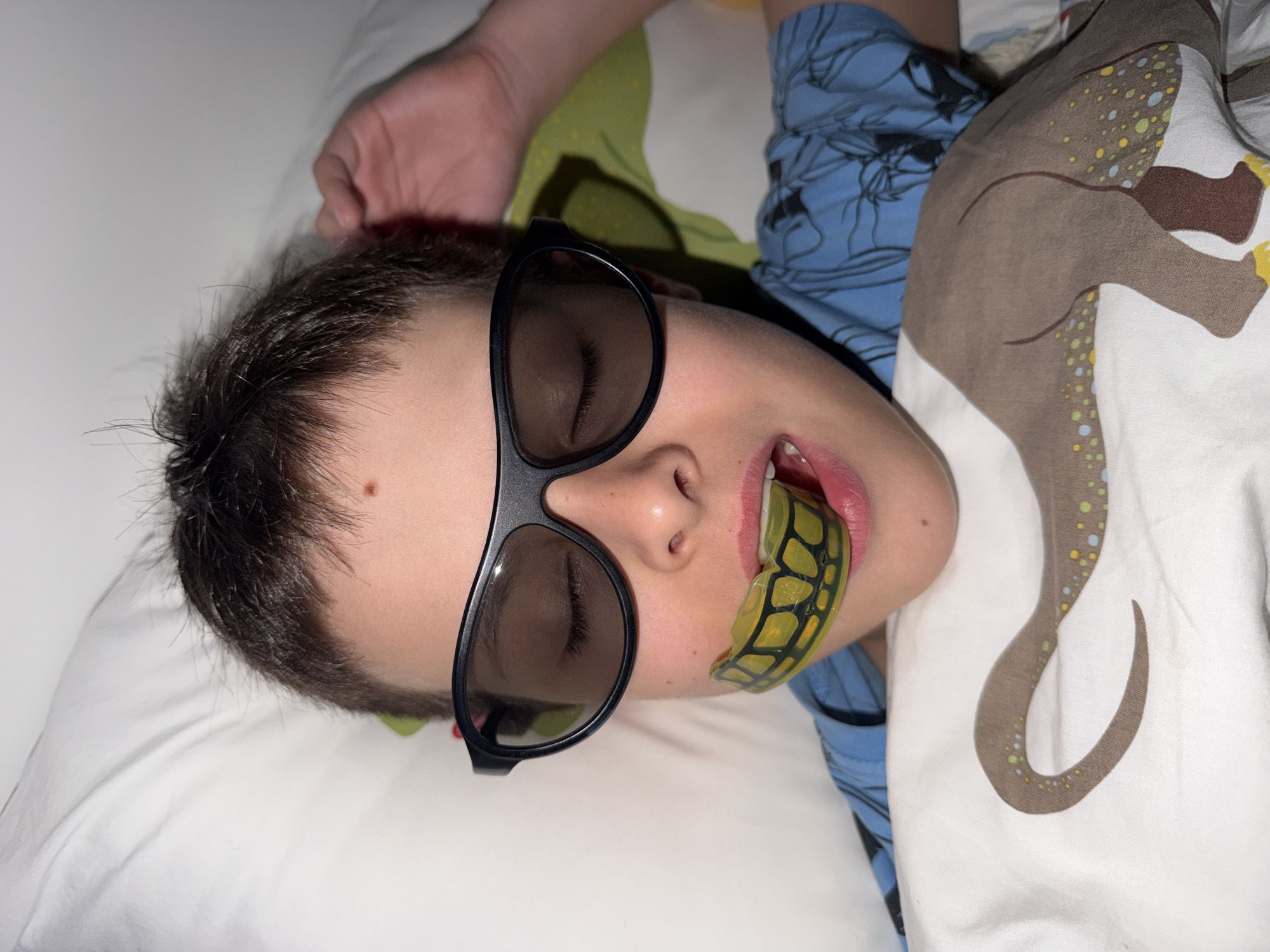 A medium close-up, vertically oriented image, shows a child lying on a white pillow, likely in a bed. The child, who appears to be a boy, is positioned with his head central in the frame, facing slightly upwards with his mouth slightly open. He has dark, short hair.
He is wearing black-framed sunglasses, beneath which his eyes are closed. A prominent feature is a yellow mouthguard with a black grid pattern, partially inserted in his mouth. His lips are pink and slightly parted.
His right hand is visible resting on the white pillow above his head. He is dressed in a light blue long-sleeved shirt with a darker blue pattern. A white duvet cover or sheet with a distinctive brown animal print, possibly a dinosaur or dragon, adorned with yellow, blue, and green speckles, covers his torso on the right side of the frame. The immediate background behind his head is a plain white surface, likely another pillow or wall.
The scene suggests the child is resting or asleep, possibly after watching a movie, given the sunglasses, or engaged in imaginative play. The lighting is soft and diffused, indicating an indoor setting, perhaps a bedroom, with no visible external light sources. There is no visible text in the image. No specific visual cues indicate the location as Votkinsk, Russia.