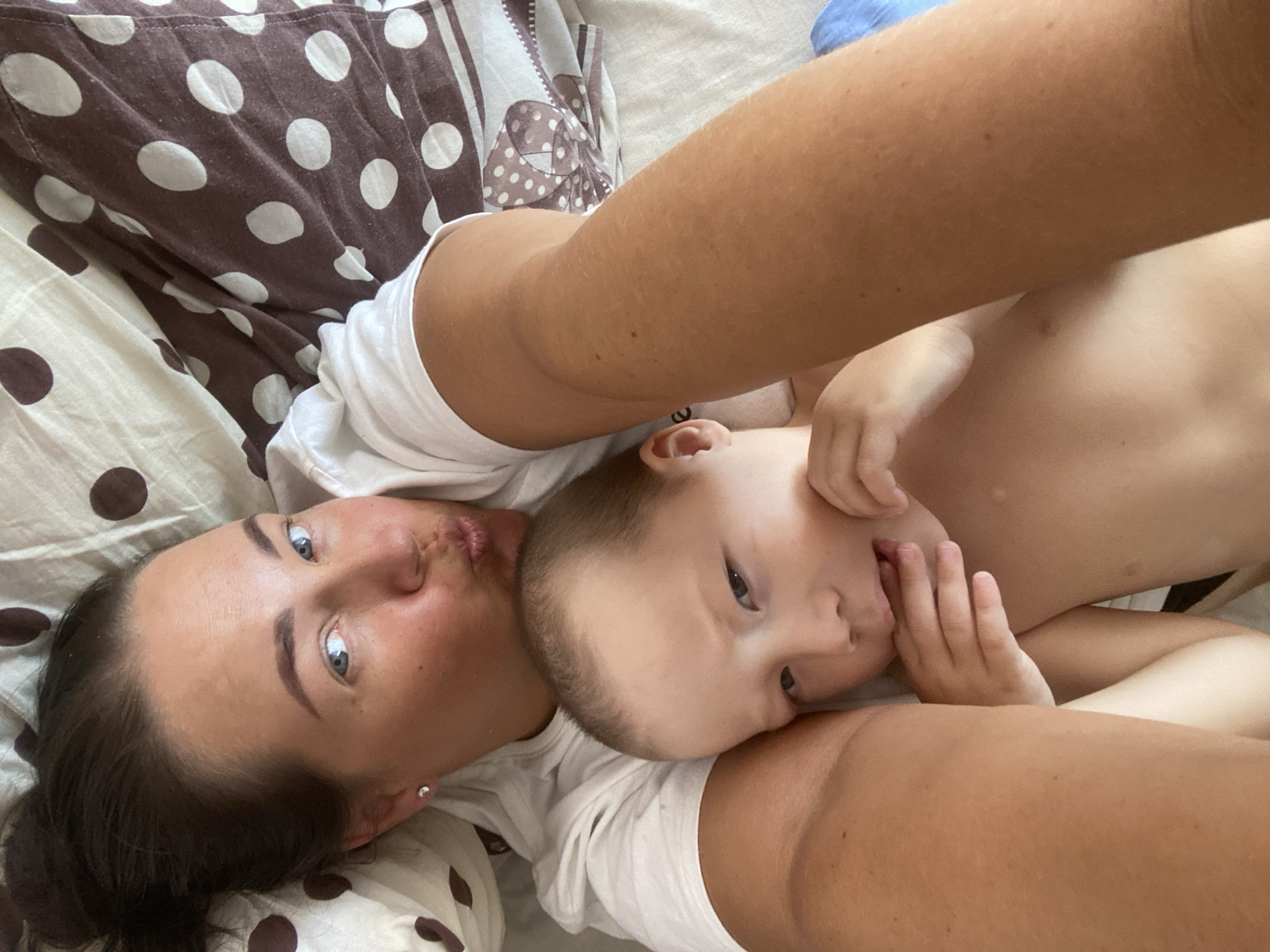 The image depicts a close-up, overhead view of a woman and a young child lying down on a bed. The woman is on the left, her face turned towards the camera with her lips pursed. She has blue eyes and dark hair pulled back. She is wearing a white t-shirt. The child is on the right, lying on their side with their head resting near the woman's face. The child has short, light brown hair and is also wearing a white t-shirt. The child's hand is near their mouth.
The bedding visible in the image has a brown and white polka dot pattern. The overall impression is of a relaxed, intimate moment between the two individuals. The lighting suggests it could be daytime, but no specific time can be determined. There are no discernible external location cues such as windows or distinct furniture that would indicate a specific room or outdoor setting beyond being indoors on a bed. The emotions conveyed are warmth and affection. No visible text is present in the image. The context of Sochi, Russia, is provided but not evident from the visual content of the image itself.