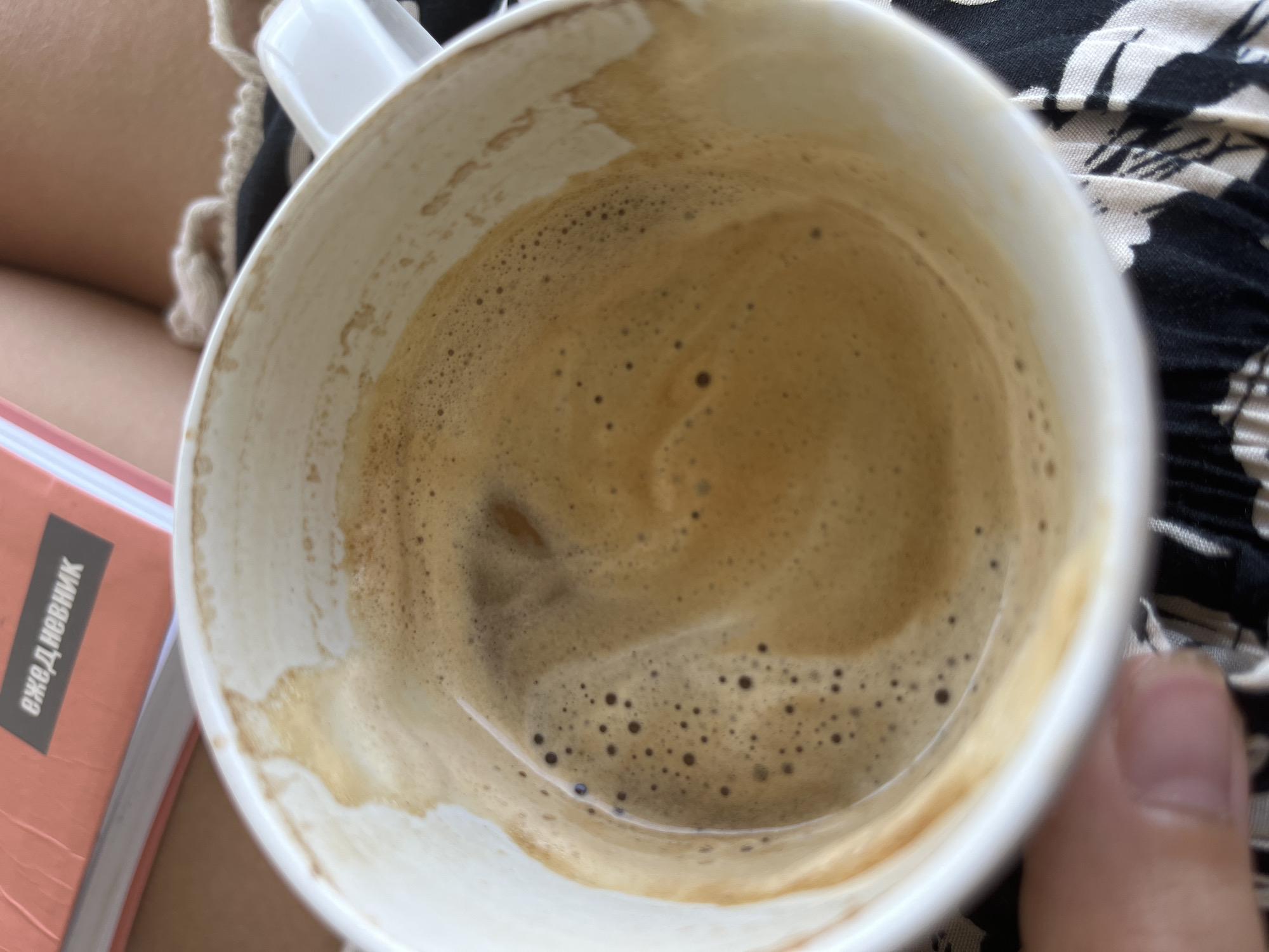 The image shows a partially consumed cup of coffee with visible coffee residue clinging to the inside. The cup is white with a handle. A portion of a notebook with Russian text, "ЕЖЕДНЕВНИК" (diary), is visible on the left. The background includes a person's lap with clothing that has a black and white pattern. A thumb is visible on the bottom right. The environment and the city of Baan Lamai, Thailand cannot be determined from the image.