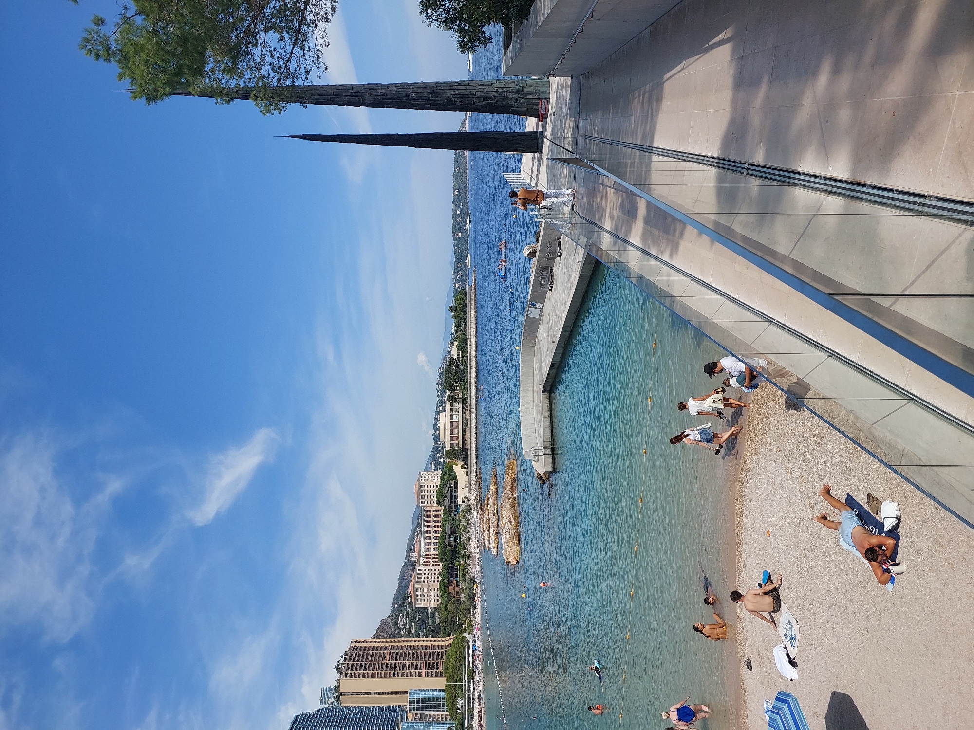 The image depicts a sunny day at a beach in Monaco, with the Mediterranean Sea in the background. The scene shows a beach with several people relaxing, swimming, and walking. There are buildings in the background, including high-rise apartments and hotels, characteristic of the Monaco skyline. The sea is calm and blue, with some boats visible in the distance. There is also a modern architectural element with glass railings, which appears to be a pathway or viewing area overlooking the beach and sea. The overall atmosphere is one of leisure and vacation.