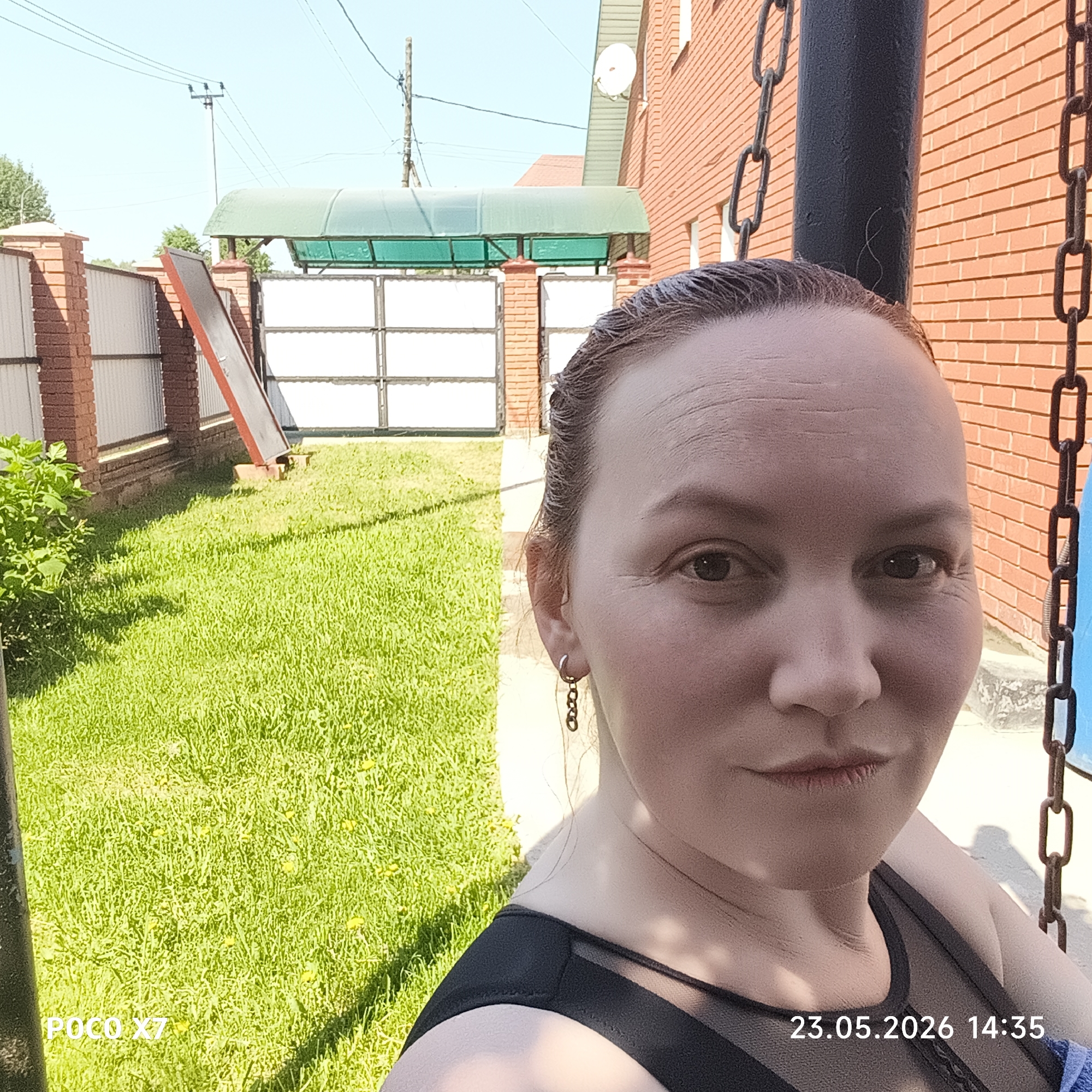 A medium outdoor selfie of a Caucasian woman with light skin, taken on May 23, 2026 at 14:35 (2:35 PM). The woman is seated on a swing, with a chain visible on the right side of the frame. She wears a dark sleeveless top. Her hair is pulled back. She is wearing drop earrings.
The background includes a green lawn, a brick house, and a metal gate with a green awning. A fence runs along the left side of the lawn.
The image metadata includes the "POCO X7" model designation. This image was taken in the city of Chaikovsky, Russia.