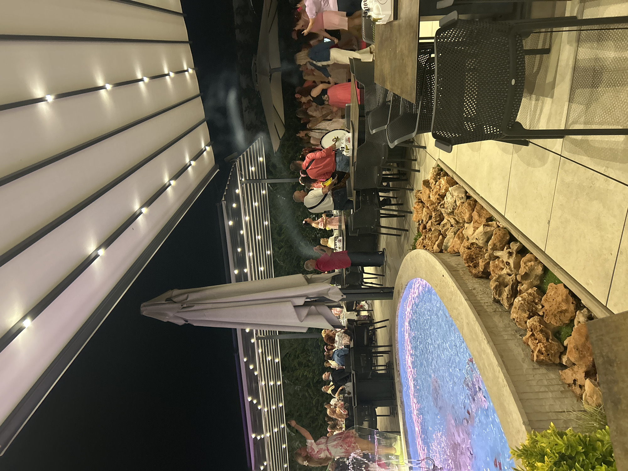 The image shows an outdoor dining area at night. Several people are seated at tables enjoying a meal, with a musician or performer entertaining them. A fountain or water feature is visible to the right, surrounded by rocks. The area is illuminated by string lights under an awning. The tables are set with chairs, and the overall atmosphere suggests a festive or celebratory occasion. The location is Kazanlak, Bulgaria.