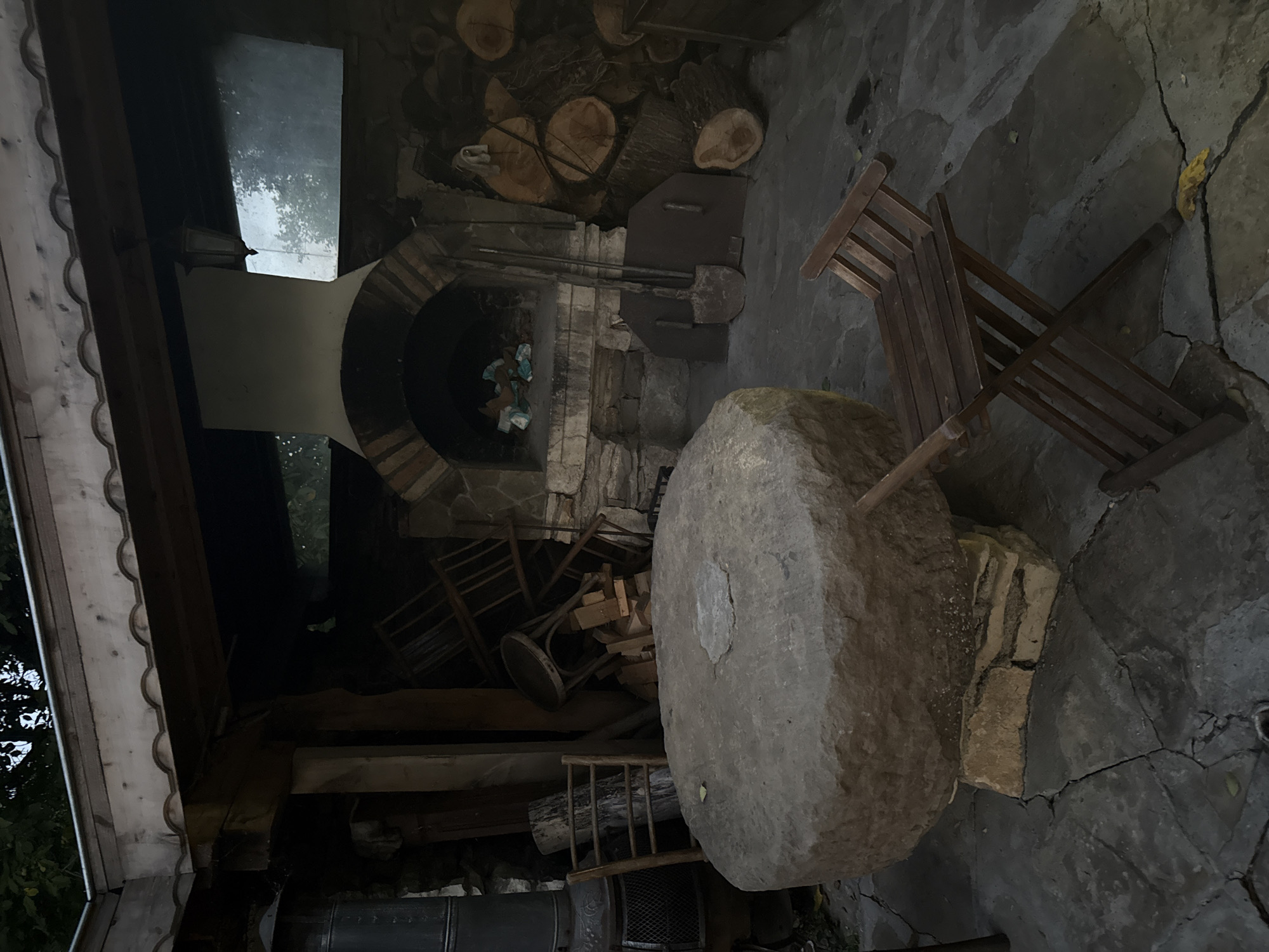 This image depicts an outdoor patio area at dusk, likely in Petrevene, Bulgaria. The scene features a large, rough-hewn stone table dominating the foreground, supported by stone blocks. To the right of the table, a wooden folding chair is positioned on a cracked stone patio. In the background, a brick-built oven with an arched opening is visible, containing some colorful debris. Adjacent to the oven is a stack of firewood. To the left of the oven, a metal stovepipe extends upwards, and behind it, several wooden chairs and miscellaneous wooden items are stored. The area is covered by a wooden roof structure with decorative trim. The overall impression is one of a rustic, somewhat cluttered outdoor space.