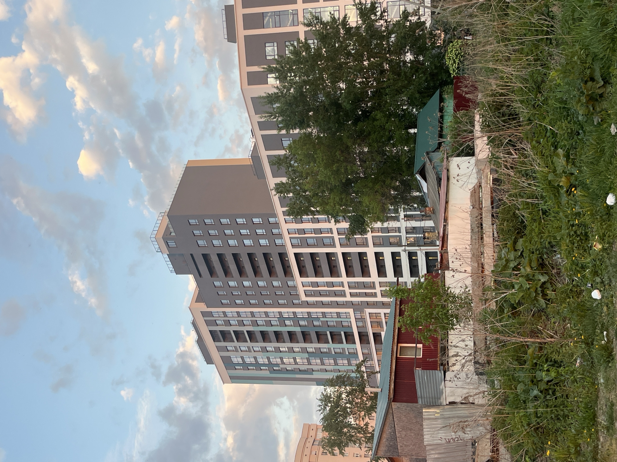 The image shows a tall, modern apartment building in Yekaterinburg, Russia. The building has a gray and white facade with many windows. In the foreground, there's a stark contrast: a dilapidated area with overgrown vegetation and old, run-down structures. The sky is partly cloudy. The scene depicts the juxtaposition of modern urban development and decaying infrastructure in a specific urban context.