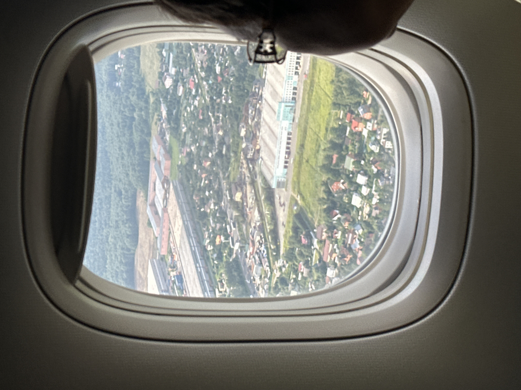 An aerial view through an airplane window captures a suburban area with a mix of industrial buildings and residential houses. The foreground shows the edge of the airplane's interior, with the curved window frame partially obscuring the scene. A person's head and shoulder, wearing glasses, are visible on the right side of the frame, looking out the window. Below the window, a long, light-blue industrial building with a white roof dominates the middle ground, surrounded by green fields and smaller buildings. Further in the distance, a dense forest forms the backdrop, with a strip of industrial development featuring long, red-roofed buildings to the left of the main industrial structure. The scene suggests a location on the outskirts of a city, likely in an area that combines industrial activity with residential development. The view corresponds to the city of Nochvino, Russia, characterized by such mixed-use landscapes.