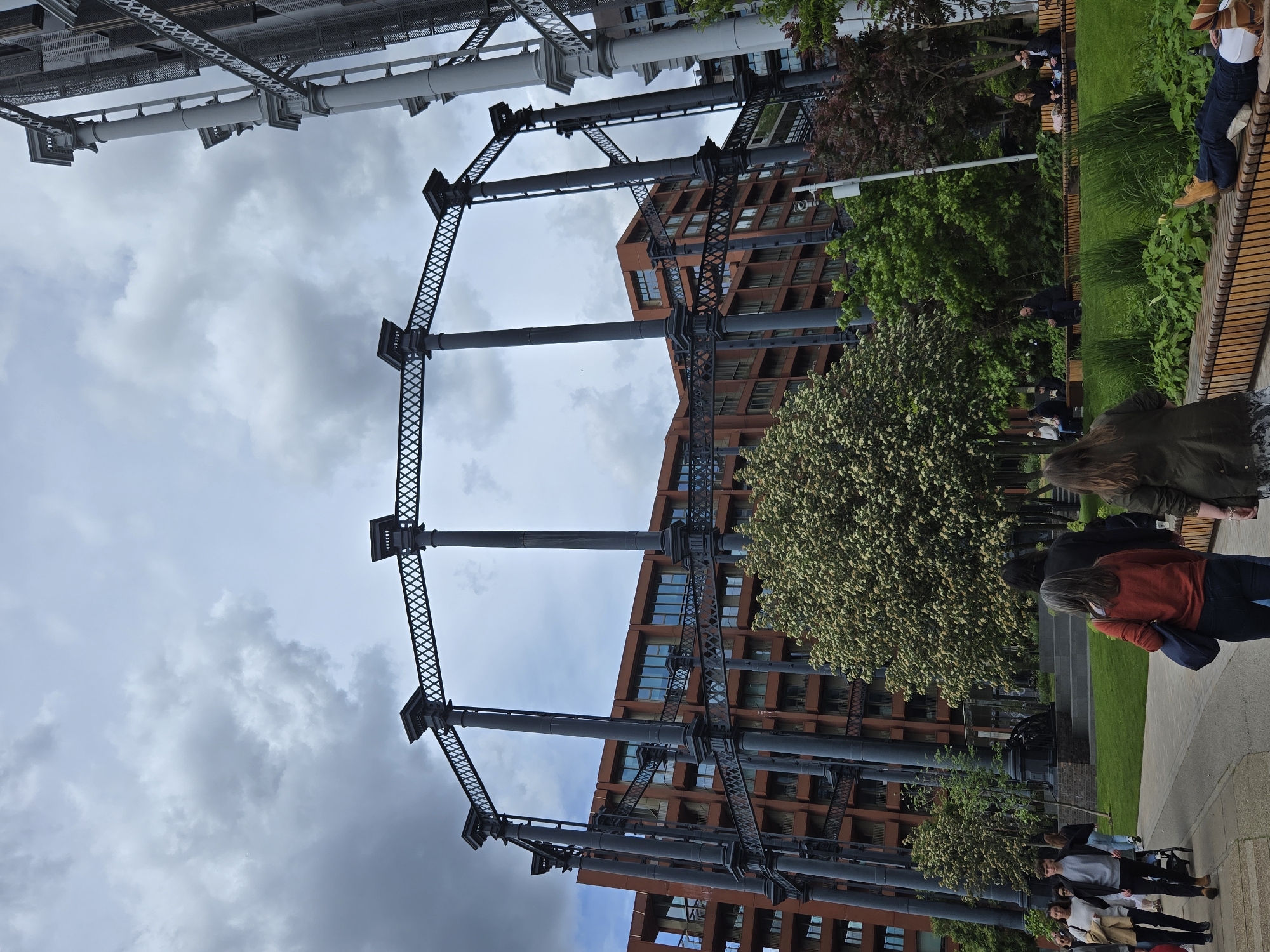 The image depicts the Gasholders Park apartments in King's Cross, London, United Kingdom. The scene includes the refurbished cast iron frames of former gas holders, now incorporated into the architecture of modern apartment buildings. Several people are present, including individuals walking and others seated on benches in the park area. The environment includes trees, grass, and paved pathways. The sky is cloudy.