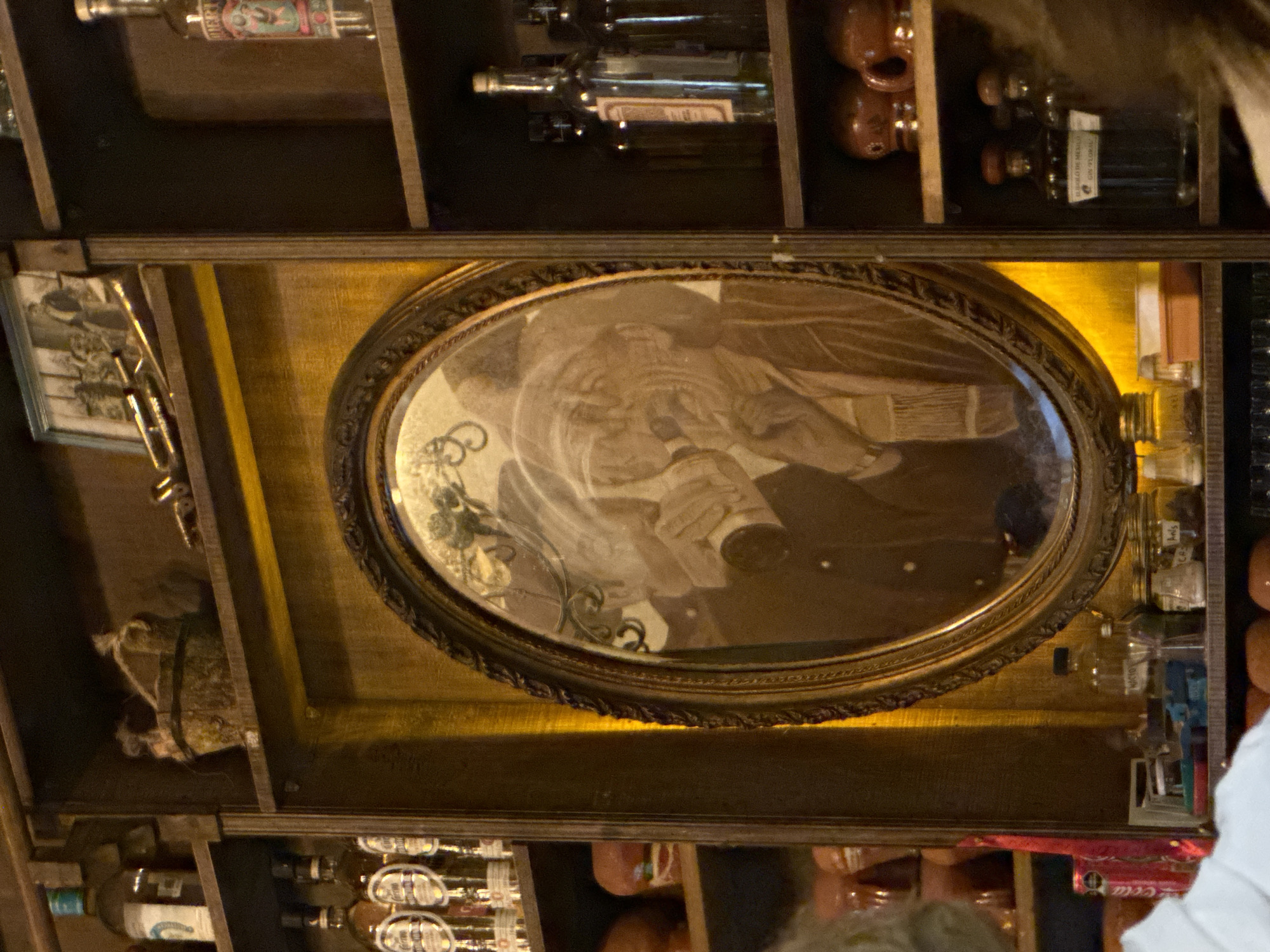 This image is of a shelf in what appears to be a bar or restaurant, likely in Puebla, Mexico, given the presence of Mexican spirits and decorations. The focus is on an oval-shaped mirror mounted within a wooden frame in the center of the shelf. The reflection in the mirror is blurry, but seems to be depicting a man wearing a jacket or suit.
The shelves contain a variety of items including:
- Bottles of liquor
- Ceramic figurines
- Framed photograph
- A small burlap sack
- Miscellaneous items such as small bottles and containers.
The overall lighting suggests an indoor environment with somewhat dim, warm lighting. Some of the shelves are illuminated with a yellow light source behind the frame. Portions of two people are visible at the bottom, indicating that it is taken at a bar or restaurant.