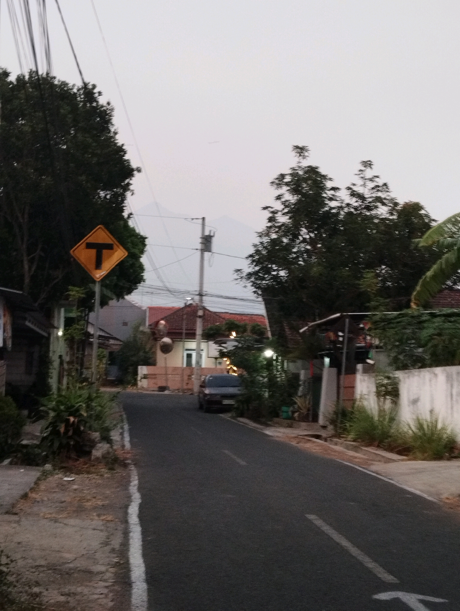 The image depicts a street scene in Salatiga, Indonesia, at dusk. A diamond-shaped yellow traffic sign with a black "T" symbol, indicating a T-intersection ahead, is prominently displayed on the left side of the road. The road surface is asphalt, with a white line marking the center or lane separation. A dark-colored car is parked on the right side of the road, partially visible. The surroundings feature houses with red-tiled roofs and some vegetation, including trees and bushes. Electrical wires are visible overhead, strung between poles. The sky is overcast, suggesting either early morning or late evening. The overall impression is a quiet, residential street in a suburban or semi-urban area.