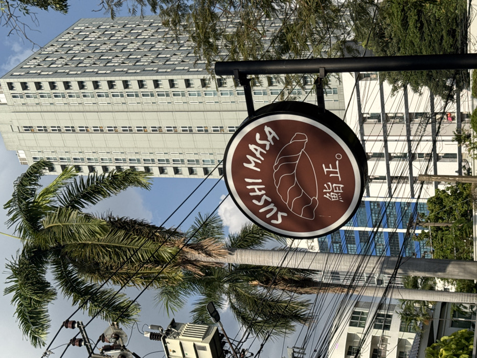 The image depicts an urban street scene in Bangkok, Thailand, centered on a circular brown business sign. The sign, edged in white, displays "SUSHI MASA" in white capital letters curved at the top, a stylized white line drawing of a sushi piece (nigiri), and two Japanese characters "鮨正" below. It is mounted on a black metal pole extending into the frame from the lower right.
In the mid-ground, a tall palm tree with green fronds is visible behind and to the left of the sign, with a second palm tree partially visible further left. Numerous black electrical wires crisscross the upper left portion of the frame, connecting to a utility pole structure on the far left that features an electrical transformer box and a street light fixture.
The background is dominated by a tall, modern high-rise building, light gray in color, with multiple rows of rectangular windows, positioned on the right side of the frame. Another building with blue-tinted windows is partially visible behind the sushi sign. Green foliage is scattered throughout the lower and mid-ground. The sky above is clear blue with sparse white clouds. No individuals are present in the scene.