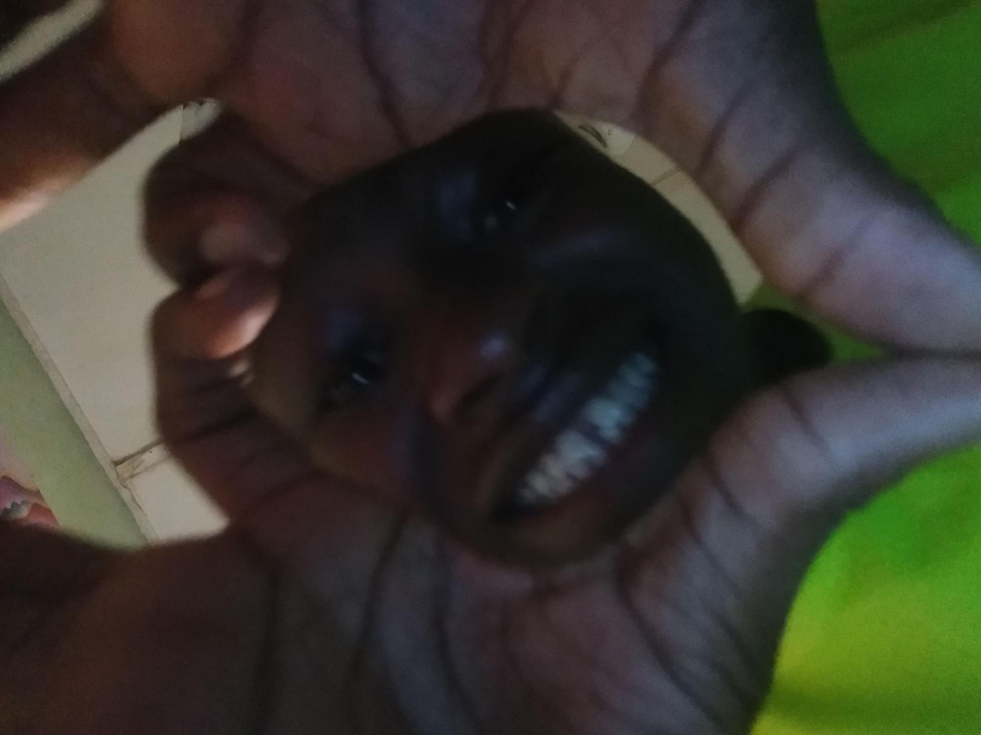 A person is holding a dark-skinned child's head in their hands, framing the face. The child is smiling, revealing their teeth. The background includes a green surface and what appears to be a wall. The location is Unknown, Unknown.