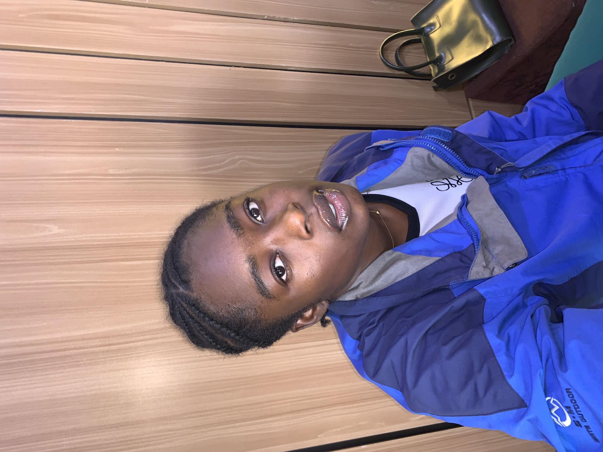 A young Black female with braided hair is looking at the camera. She is wearing a blue and gray jacket over a white t-shirt with a black collar. There is a small gold necklace around her neck. To the right of the woman, there is a dark green handbag resting on a brown object. The background consists of light brown wooden panels. The scene appears to be indoors. The location is in the city of Gura Top, Nigeria.
