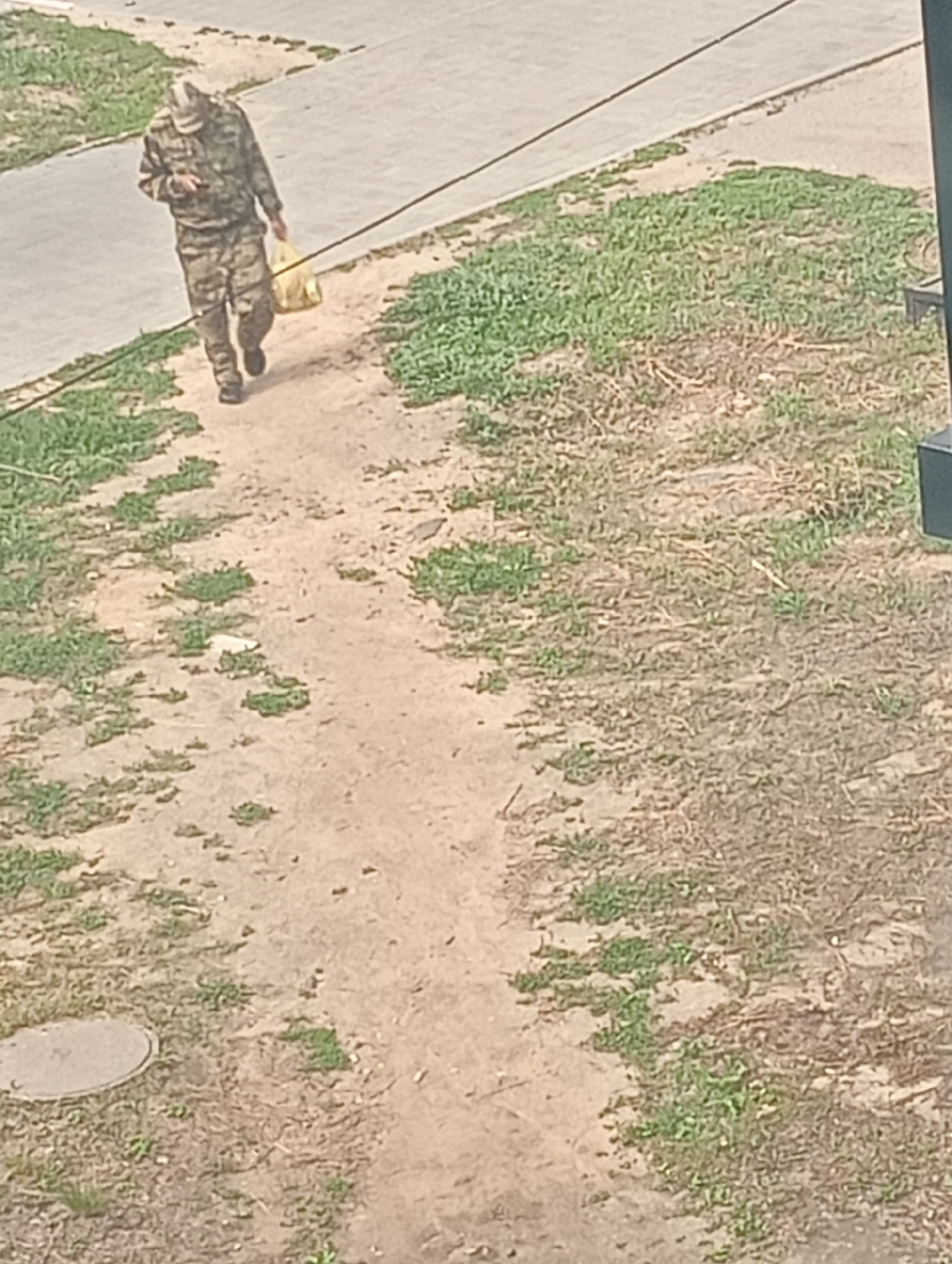 In an outdoor scene in an unknown city, a person wearing a camouflage uniform and a cap walks along a dirt path beside a paved area. The person is carrying a yellow bag in their right hand. The ground is a mixture of dirt and sparse green vegetation. A dark line runs diagonally across the upper portion of the image, likely a wire or cable. A dark rectangular object is partially visible in the far right of the frame. A round, light-colored manhole cover is visible on the dirt path in the lower left corner.