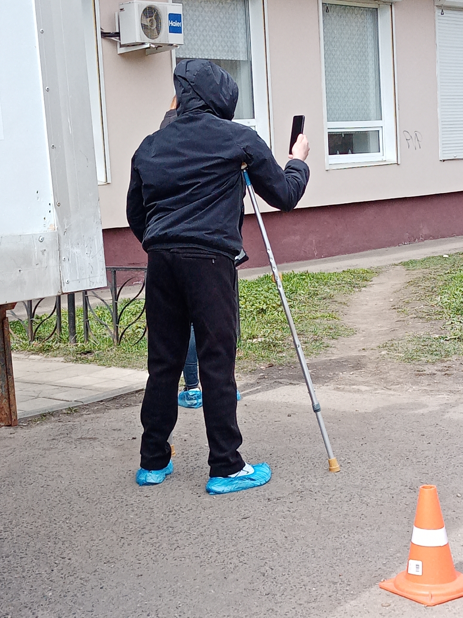 The image captures an outdoor daytime scene in Elektrostal, Russia. A person, viewed from behind, is the primary subject. They are wearing a black hooded jacket, black pants, and a bright blue disposable shoe cover over their left foot; another blue shoe cover is visible on their right foot. This person is holding a smartphone in their right hand, pointing it forward as if taking a photo or recording. They are also utilizing a single crutch, positioned next to their right side.
Another person, mostly obscured, is partially visible behind the main subject, also wearing blue shoe covers. The ground beneath them consists of asphalt, with a dirt path leading to a grassy area on the right, bordered by a low, ornate metal fence.
In the background, a light-colored building features multiple windows, some with internal blinds or external roller shutters. An outdoor air conditioner unit, clearly labeled "Haier," is mounted on the building's wall. Faint handwritten or graffiti text, "PP," is visible on the building's exterior near one of the windows. An orange traffic cone with a white reflective stripe stands on the asphalt in the foreground to the right. The lighting suggests an overcast day.