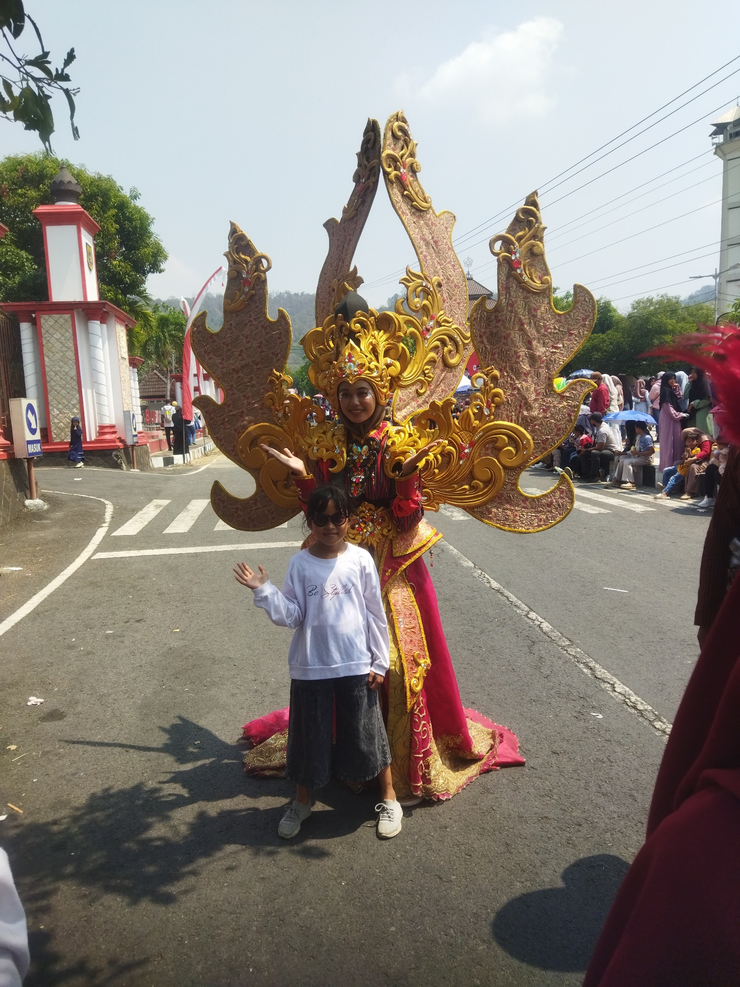 In Wonogiri, Indonesia, a young girl in a white long-sleeved shirt and dark pants stands on a paved road next to an adult in an elaborate, golden and red costume with large, ornate wings. The adult, wearing a golden headdress and makeup, is smiling and gesturing with their left hand towards the girl. The girl is also smiling and waving with her right hand. Spectators are visible in the background, some sitting and others standing, with some holding umbrellas. Buildings and trees line the street, and a white and red structure with a dome is visible on the left side of the image. The scene appears to be a street event or parade on a clear, sunny day.