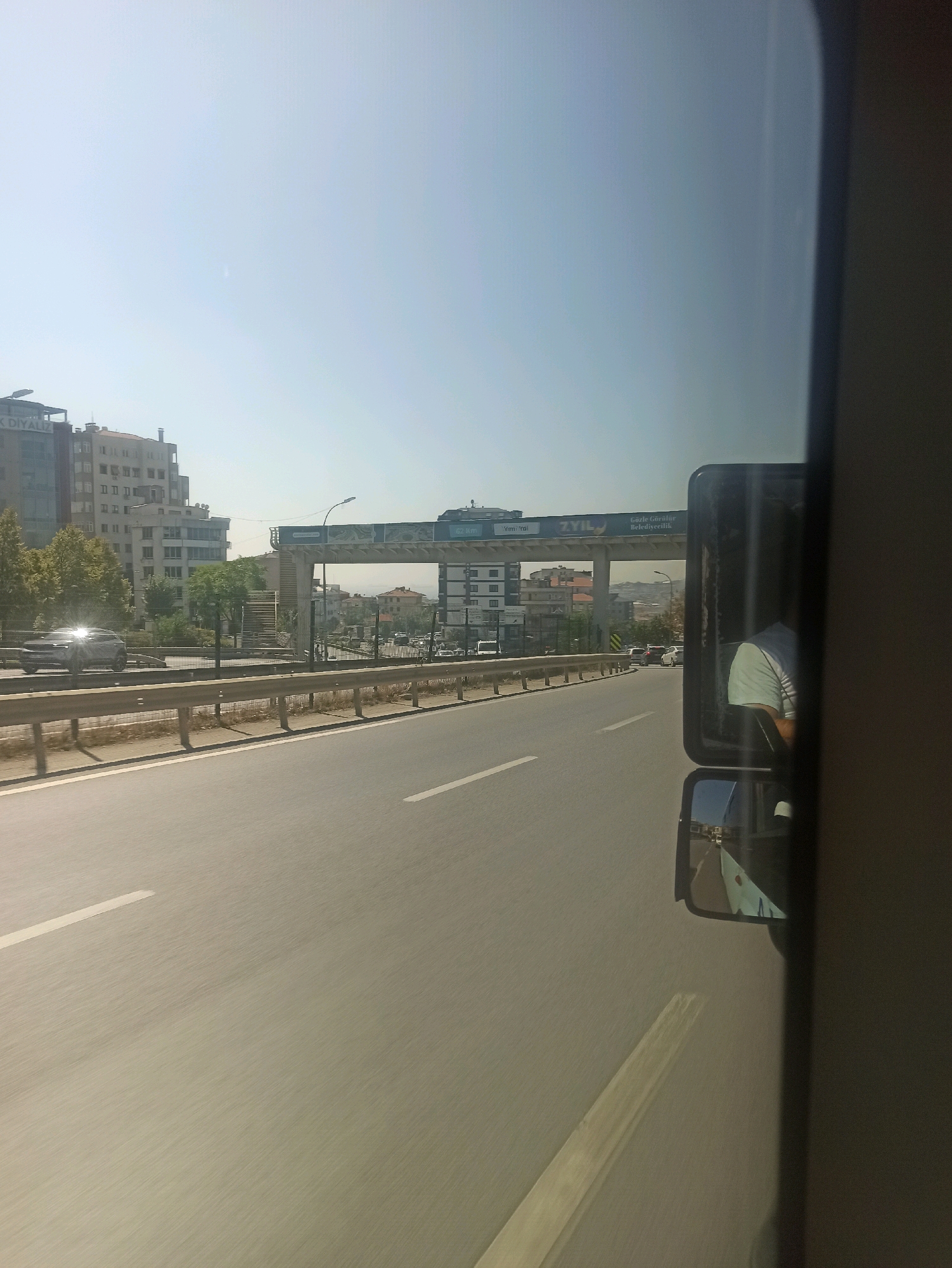 This is a photograph taken from a vehicle traveling on a highway in Pendik, Turkey. The image captures a view of the road ahead, with buildings and an overpass visible in the background. The sky is clear and bright, suggesting a sunny day. On the right side of the image, a side-view mirror reflects the interior of the vehicle and a person wearing a light-colored shirt, likely the driver. The road appears to have multiple lanes, and there is some traffic visible in the distance. The overall impression is of a typical urban highway scene on a clear day.