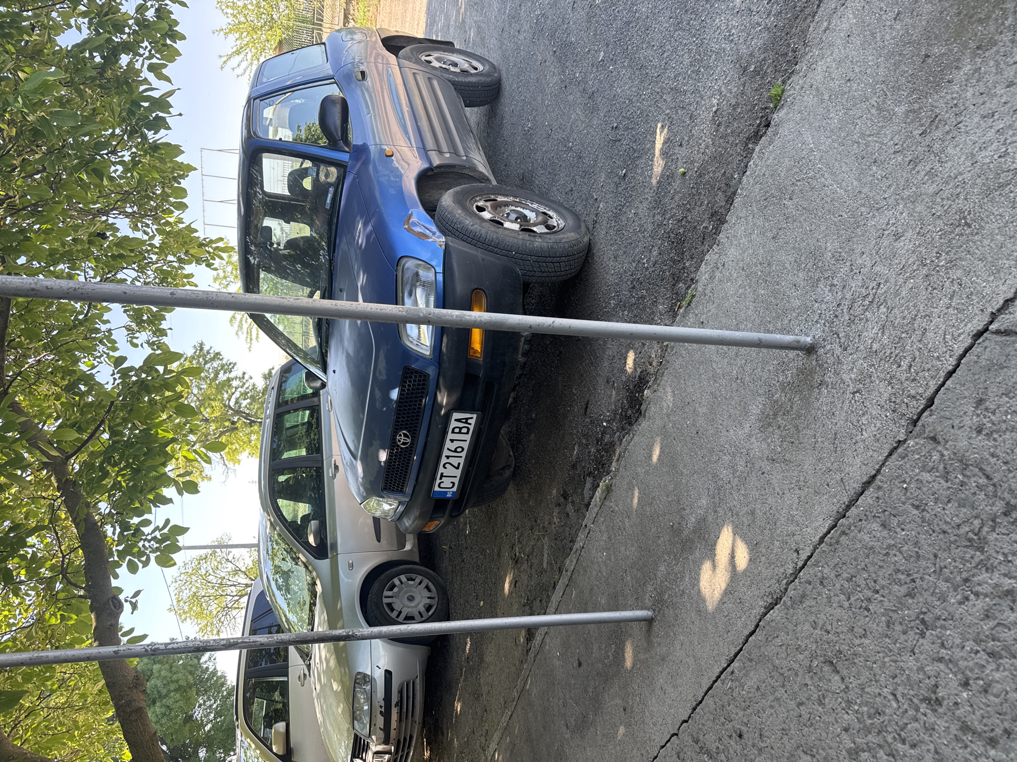 The image shows a street scene in Sarnevo, Bulgaria. Two parked cars are visible, a silver one behind a blue Toyota RAV4. The Toyota RAV4 has the license plate CT2161BA. The cars are parked on the side of the road next to a concrete sidewalk. Metal poles are present, seemingly part of a barrier or railing. A tree with green leaves is visible on the left side of the image. The sky is visible through the leaves, indicating daylight. The road surface is asphalt. There are light spots visible on the sidewalk and road, indicating sunlight filtering through the trees. The Toyota RAV4 appears slightly damaged on the front passenger side.
