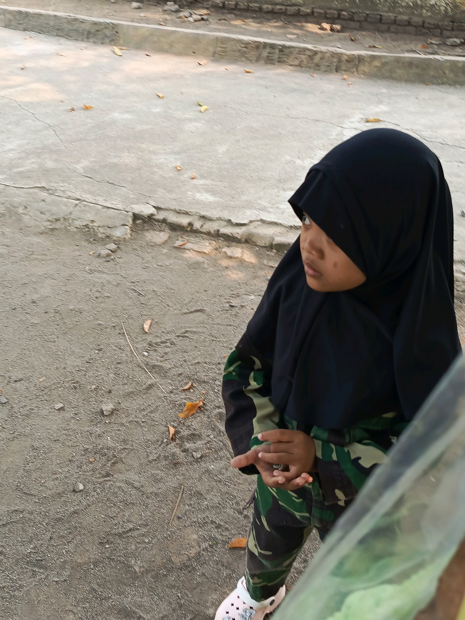 A young girl wearing a black hijab and camouflage pants stands on a gravel and concrete surface outdoors. She is looking to her right, with her hands clasped in front of her. The background shows a concrete curb and more gravel. Fallen leaves are scattered on the ground. The image appears to be taken in Karya Dadi, Indonesia.