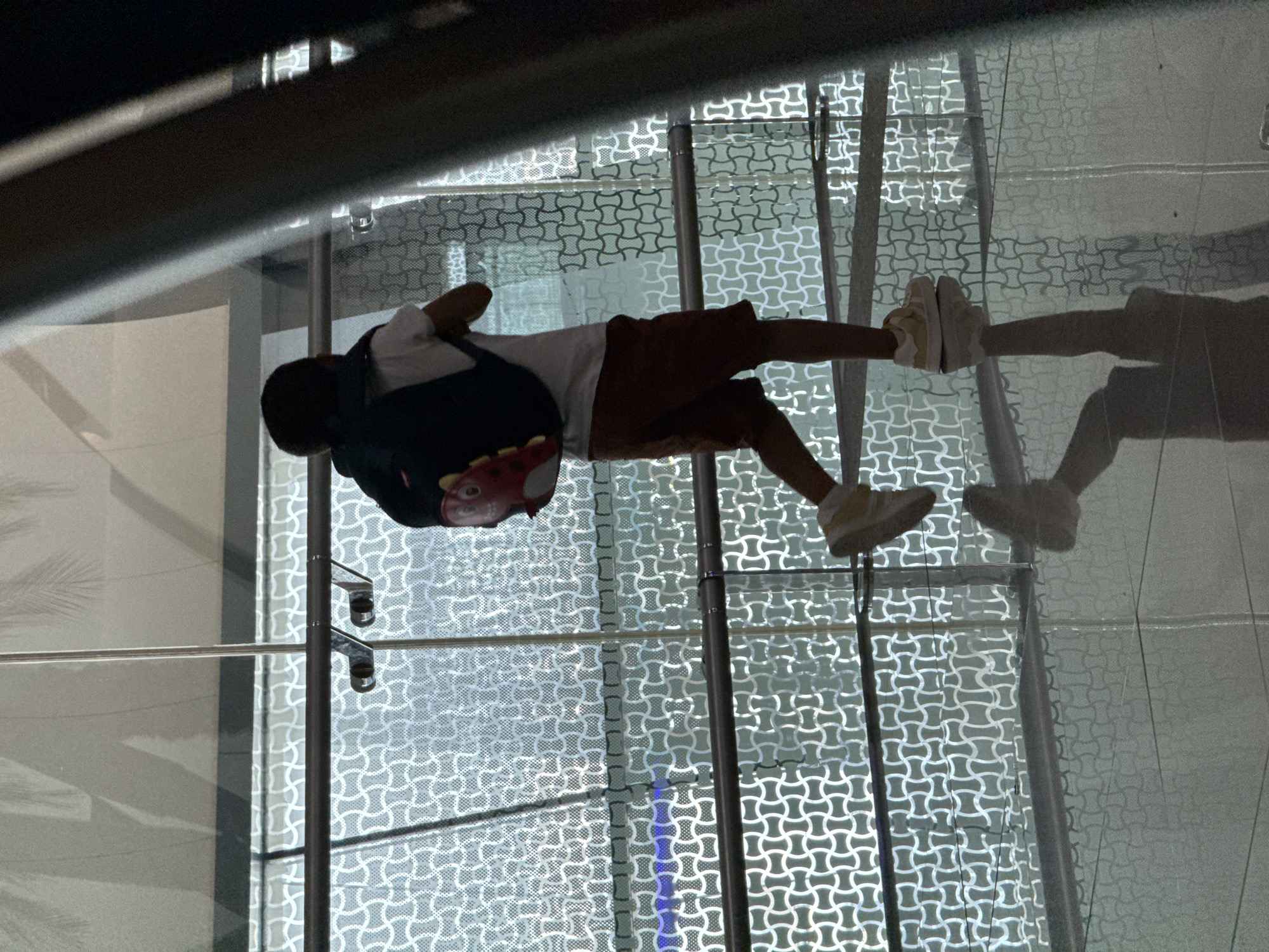 A young boy, seen from behind and in profile, walks forward while wearing a dark backpack with a red character accessory. He is wearing a white t-shirt and brown shorts, with light-colored sneakers. The boy is walking on a polished floor that reflects his image. He is in a modern indoor space, likely an airport or a large public building, with a large glass wall featuring an intricate geometric pattern. Sunlight streams through the glass, creating shadows and highlighting the textures of the environment. The scene is located in Seeb Province, Oman.