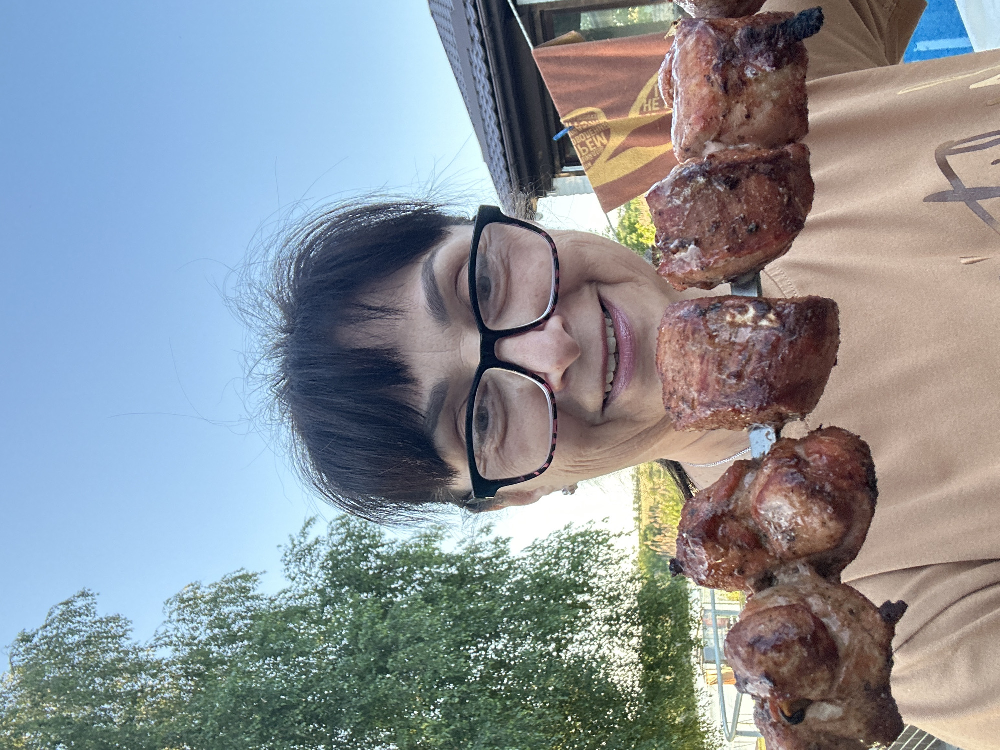 A woman with dark hair, wearing glasses, smiles at the camera while holding a skewer of cooked meat in front of her. She is outdoors, with a clear blue sky above. To her left, a green tree is visible, and to her right, a brown roof of a building and a towel hanging on a clothesline can be seen. The background also shows some vegetation and a body of water, suggesting a rural or suburban setting. The woman appears to be in Hnarovske, Ukraine.