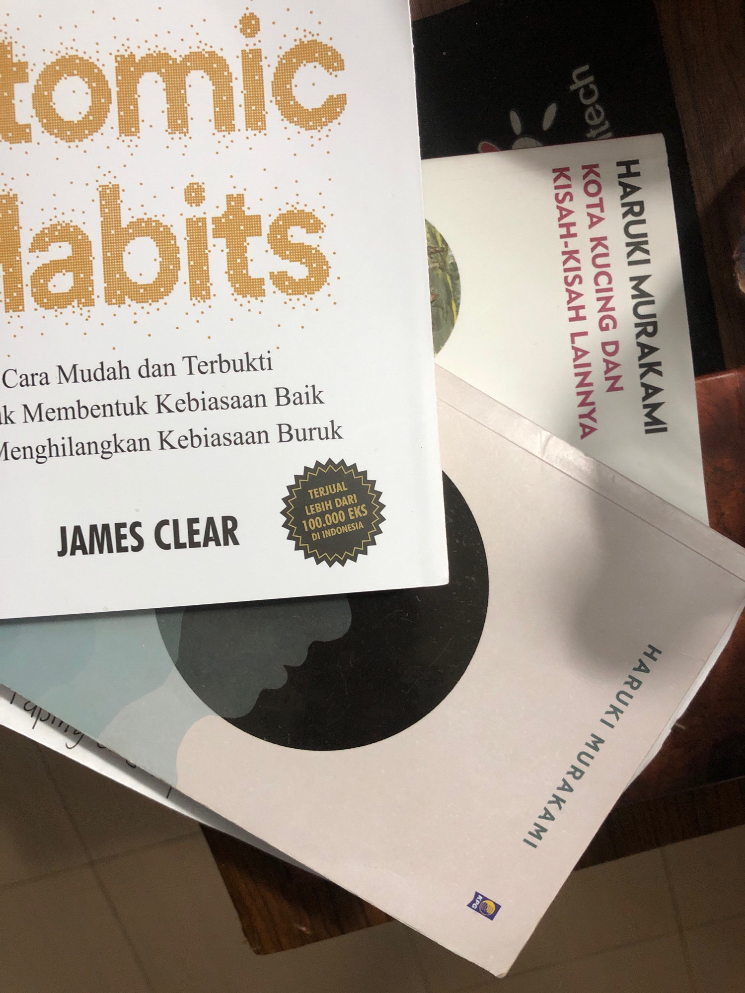 The image displays a stack of books on a wooden surface, likely a table. The top book, titled "Atomic Habits" by James Clear, is prominently featured, with its cover partially visible. Below it, another book by Haruki Murakami, identified as "KOTA KUCING DAN KISAH-KISAH LAINNYA" (The City and Its Uncertain Beasts), is also visible. A third book, also by Haruki Murakami, lies beneath the others. The books appear to be in good condition. The lighting suggests an indoor setting. The image lacks any specific visual cues that would allow for the determination of its location within Lhokseumawe, Indonesia.