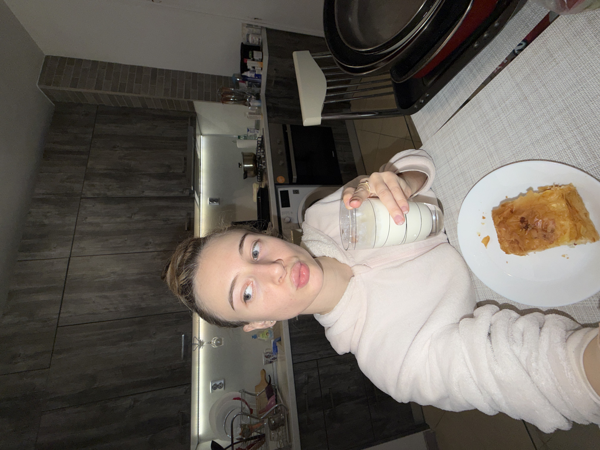The image depicts a young adult female seated in a residential kitchen, located in Plovdiv, Bulgaria. The female, with light skin and light brown hair styled in a bun, is shown from the chest up, wearing a light pink hooded fleece sweatshirt. She has a pursed-lips expression and is looking towards her left. Her right hand, featuring rings on the ring and middle fingers, holds a clear glass containing a white liquid. On the table in front of her, on a white plate resting on a light grey woven table covering, is a piece of golden-brown, flaky pastry.
The kitchen environment features dark wood-grain upper and lower cabinets, with light-colored countertops illuminated by under-cabinet strip lighting. A washing machine, an oven, and a stovetop with a pot are visible appliances. A dish drying rack filled with various kitchen items is located near a sink area. A corner wall segment is clad in brick-patterned tiles. To the right of the table, a light-colored chair and a stack of dark-colored frying pans or baking trays are present. The scene is illuminated by artificial light sources.