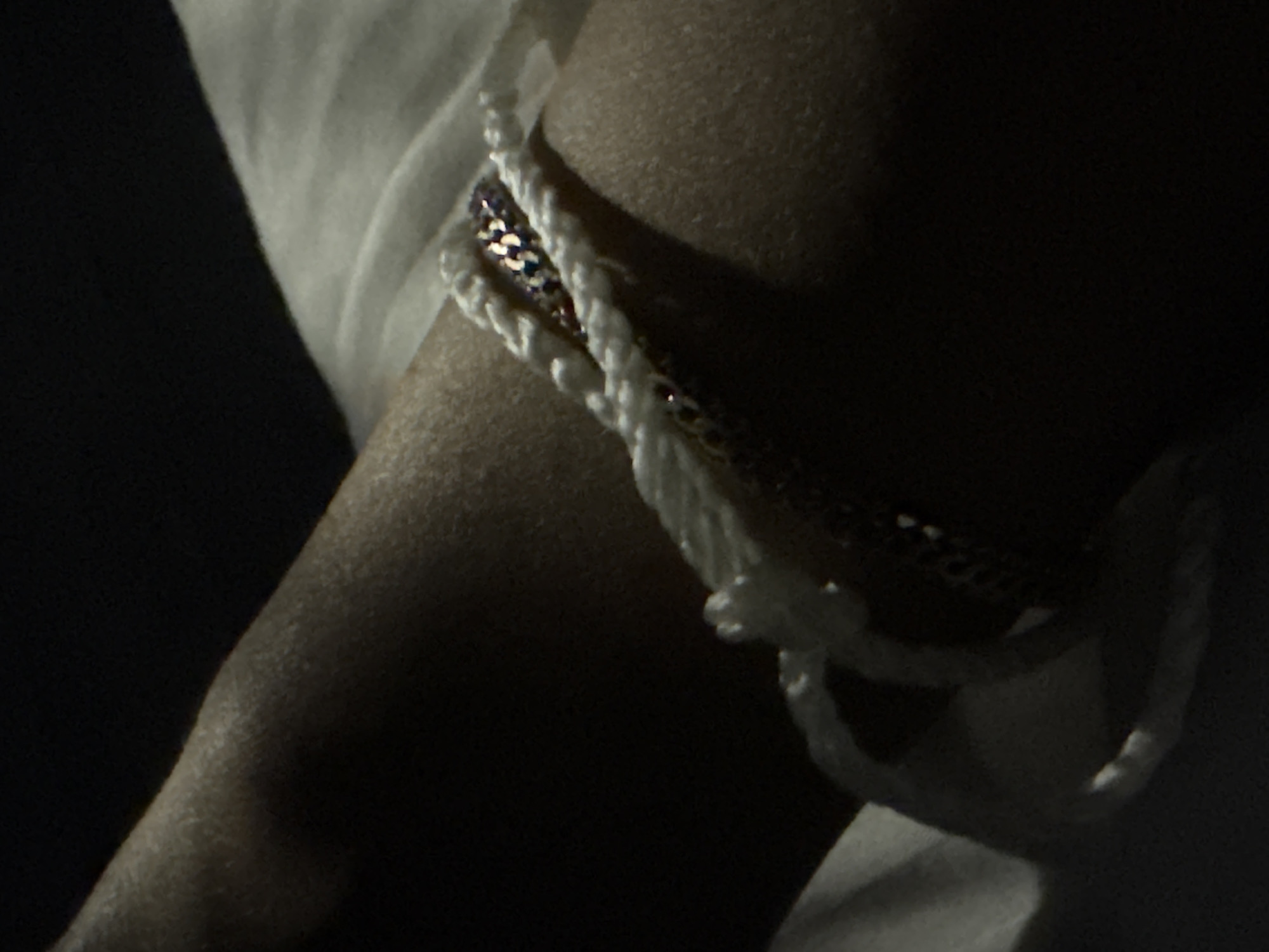 A close-up, dimly lit shot focuses on a person's arm wearing a bracelet. The bracelet appears to be made of white braided cord and a strip of shimmering, multi-colored beads, possibly sequins or rhinestones. The arm is resting on a white surface, possibly fabric. The background is dark and out of focus. The location is Doma 1096 Km, Russia.