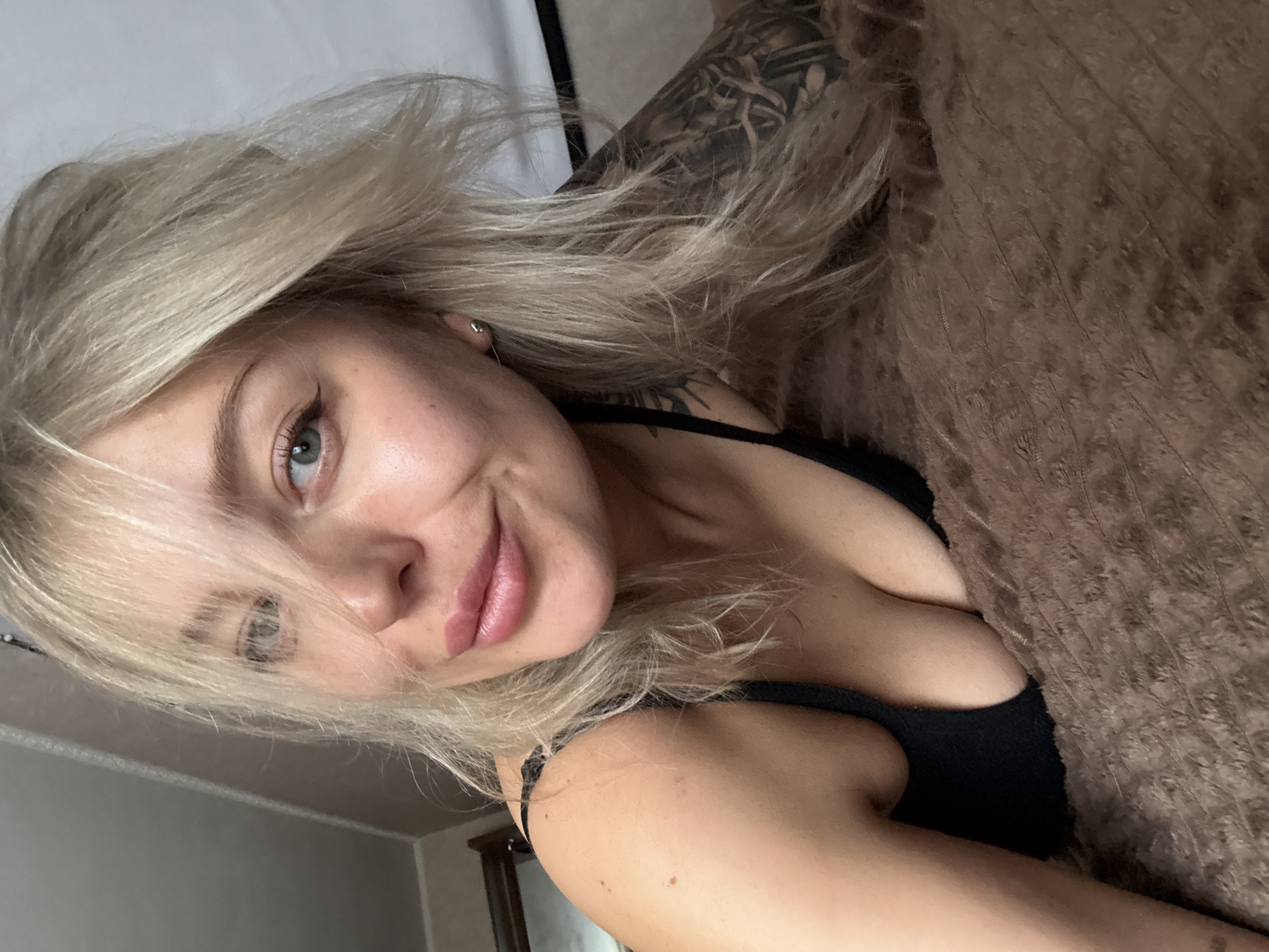 A woman with blonde hair and a tattoo on her arm is lying on a brown, textured surface, possibly a bed or couch. She is wearing a black top. The scene appears to be indoors, with a wall and doorway visible in the background. The location is potentially Borzya, Russia.