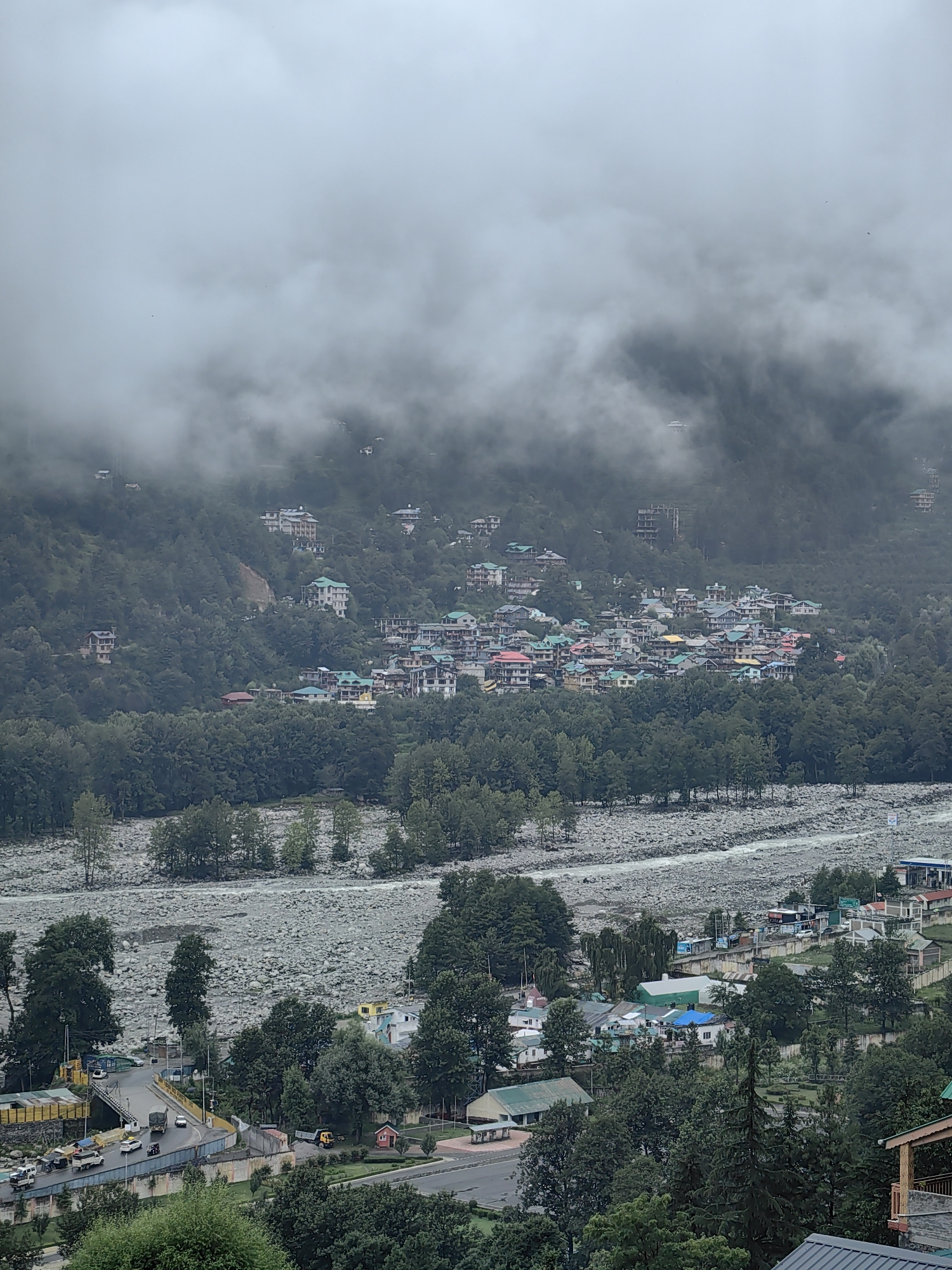 The image captures a wide, elevated view of a valley in Vashisht, India, under a cloudy, overcast sky. The scene is dominated by lush green forested hillsides dotted with numerous buildings, suggesting a densely populated area within the valley. A prominent river, appearing wide and stony, flows through the lower part of the image, with a road and bridge crossing it in the foreground. Several vehicles, including cars, trucks, and a motorcycle, are visible on the road, indicating ongoing activity. The buildings in the town are varied in architecture, with many featuring colorful roofs, some in shades of green, red, and yellow, and others with more neutral tones. The hillsides are partially obscured by thick, low-hanging clouds, creating a misty and atmospheric effect, particularly towards the upper portions of the frame. The overall impression is of a vibrant community nestled within a natural, mountainous landscape.