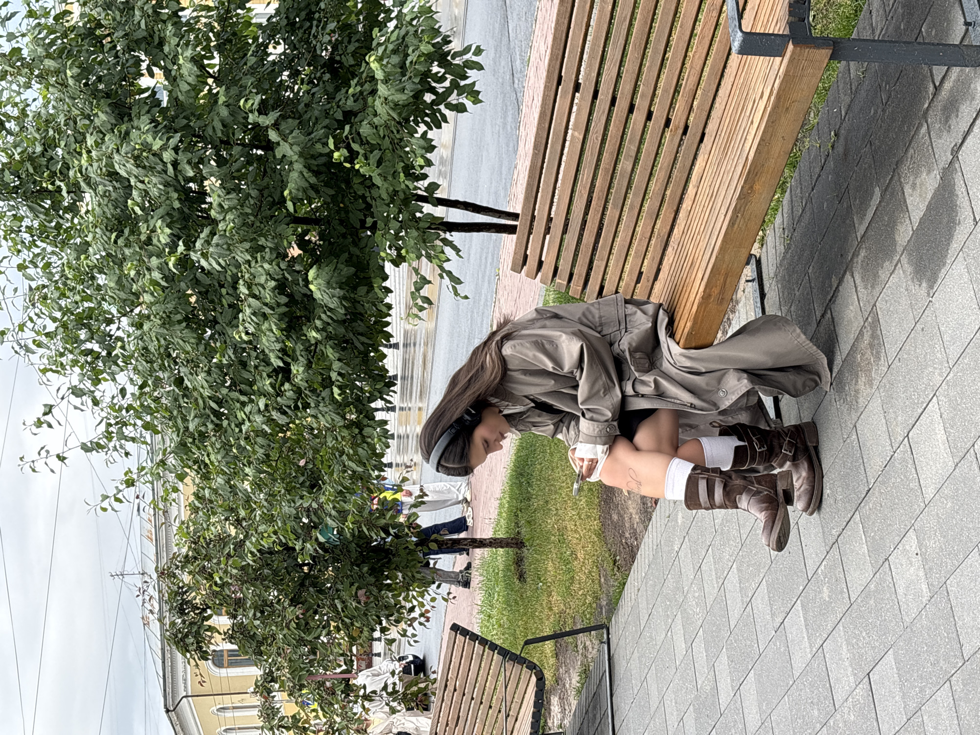 A young woman is seated on a wooden park bench, looking at her phone. She is wearing a light brown trench coat, black shorts, white socks, and brown boots. She has dark hair and is wearing headphones. The bench is on a paved walkway next to a grassy area with trees. In the background, a yellow building with arched windows can be seen. The scene appears to be in a public outdoor space, possibly a park or a street in Nizhny Novgorod, Russia. The ground is wet, suggesting recent rain.