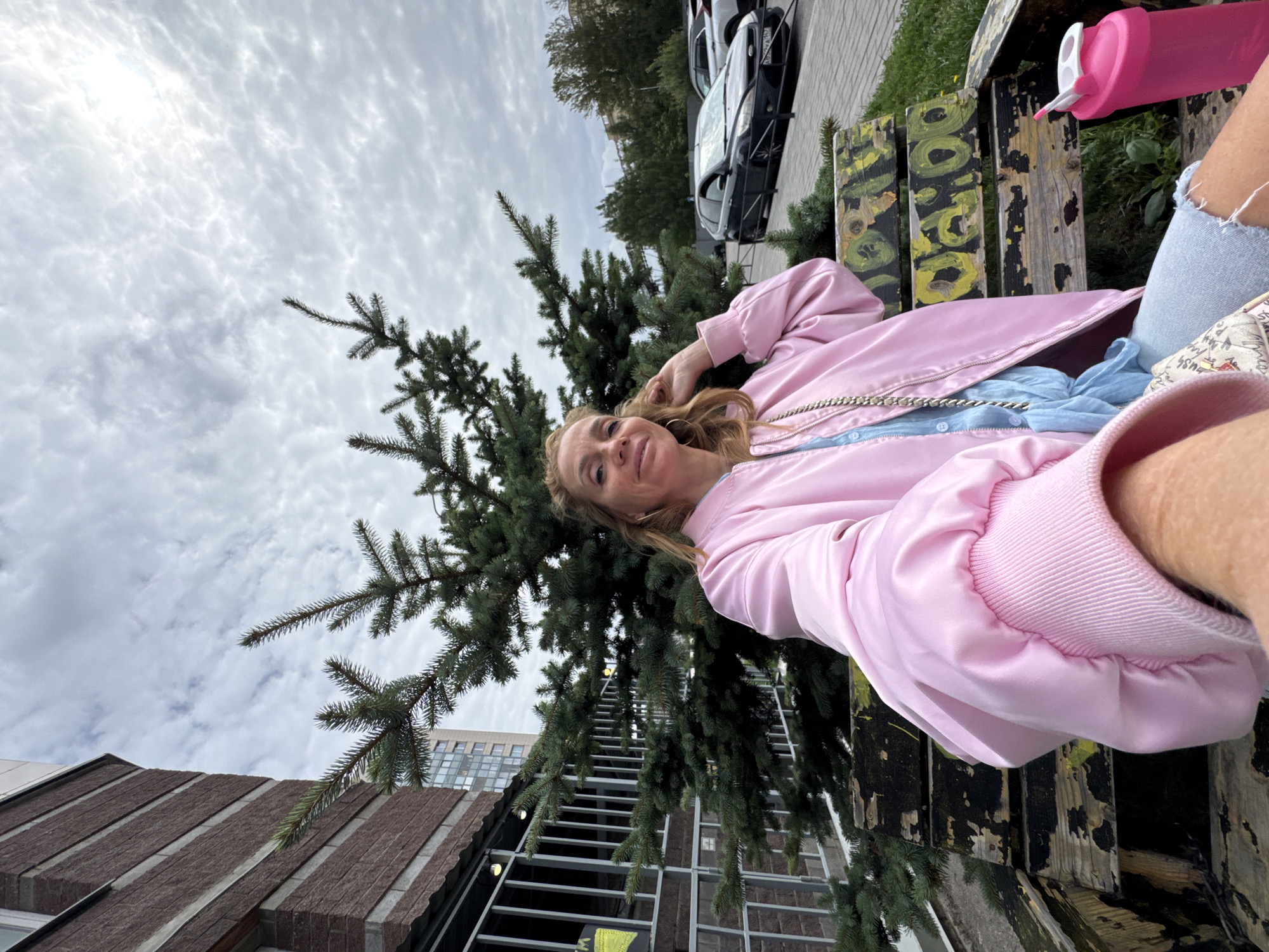 A woman with blonde hair is sitting on a wooden bench in front of a fir tree, taking a selfie. She is wearing a pink bomber jacket over a blue top and ripped denim jeans. To her right, on the bench, is a pink water bottle. In the background, a building and parked cars are visible. The image appears to be taken in the city of Sinyavino, Russia, during the day, with a cloudy sky overhead.