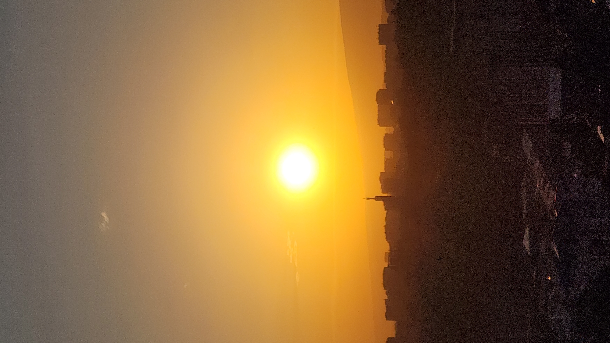 A photograph captures a city skyline silhouetted against a vibrant sunset. The sun, a bright, luminous orb, dominates the left side of the frame, casting a warm, golden-orange glow across the sky and the cityscape. The sky transitions from a pale, hazy yellow near the sun to a muted grey towards the left edge of the image.
In the distance, the shapes of buildings and possibly mountains form a dark, indistinct line against the horizon. A slender, tower-like structure is faintly visible rising from the city. The right side of the image is filled with the darker silhouettes of buildings, some appearing taller and more densely packed than others. Lights are visible within some of the structures, suggesting the city is transitioning into evening. A small, dark silhouette of an airplane is visible against the sky in the middle-right section of the image.
The scene is set during sunset, indicated by the intense color of the sun and sky. The hazy atmosphere suggests it might be a warm day or that dust or pollution is present. The overall mood is one of calm and natural beauty, with the setting sun providing a dramatic backdrop to the urban environment. The image is oriented vertically, making the sunset appear to be rising from or setting into the horizon. No text is visible in the image.