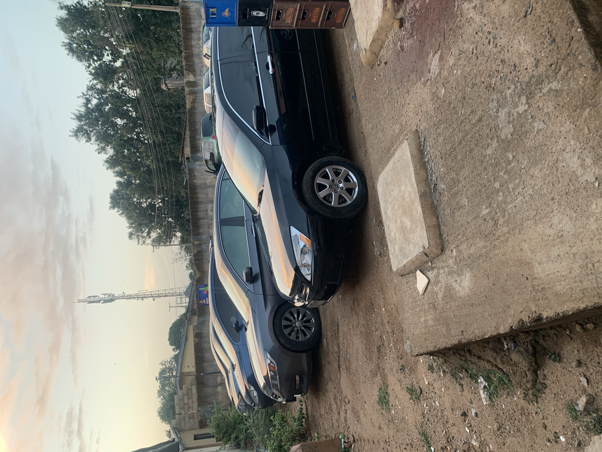 The image shows a row of parked cars along a curb in an outdoor environment, likely a roadside. The location is identified as New Stadium, Nigeria. The cars, including a black minivan, are parked closely together on a dirt and gravel surface. To the right of the vehicles is a concrete curb and sidewalk with some debris. In the background, trees, utility poles with wires, and a cell tower are visible under a hazy sky. There are crates stacked on the side of the vehicle. The lighting suggests either early morning or late afternoon. No people are visible.