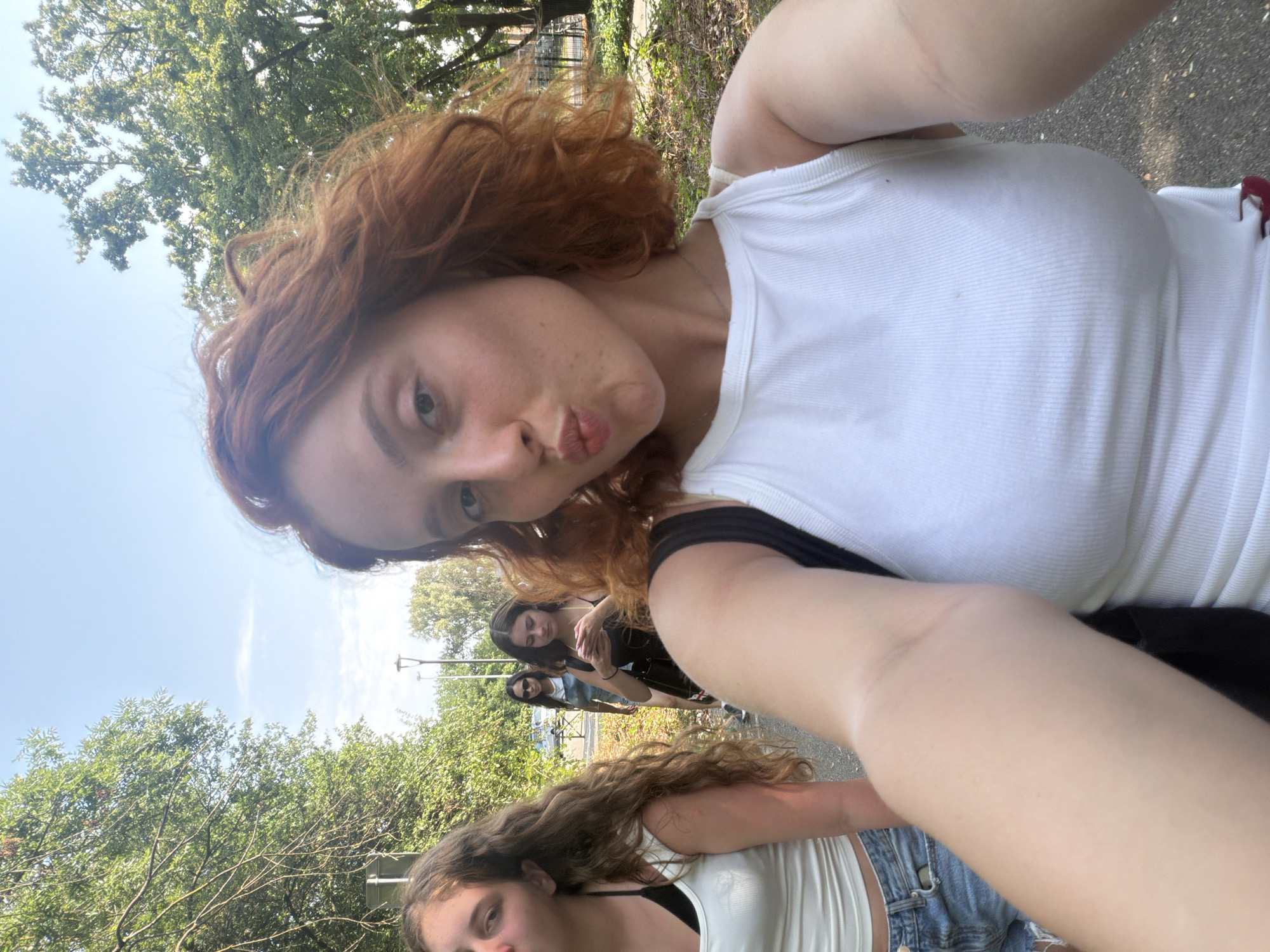 In a selfie taken outdoors, a young woman with curly auburn hair pouts towards the camera, her right arm extended to capture the shot. She is wearing a white tank top. To her left, another young woman with long, wavy brown hair looks towards the camera, wearing a white tank top and denim shorts. Behind them, two other young women walk away from the camera. The scene appears to be a park or a tree-lined path, with green trees and dappled sunlight. The location is identified as Illkirch-Graffenstaden, France.
