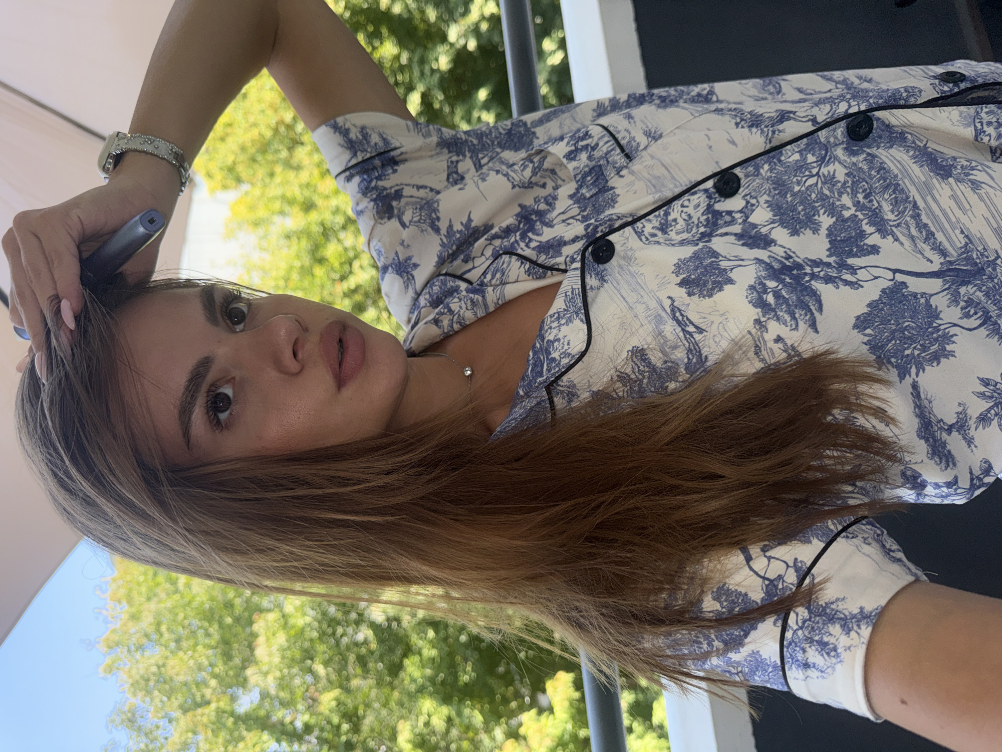 A young woman with long, auburn hair is captured from the chest up, holding a dark blue object to her head with her right hand. She is wearing a white short-sleeved pajama top with a blue toile print. A silver watch with a diamond-studded band is on her left wrist. Her left arm is raised. The background is blurred, showing green foliage and a bright sky, suggesting an outdoor setting during the day. The image does not contain any information that places the scene in Plovdiv, Bulgaria.