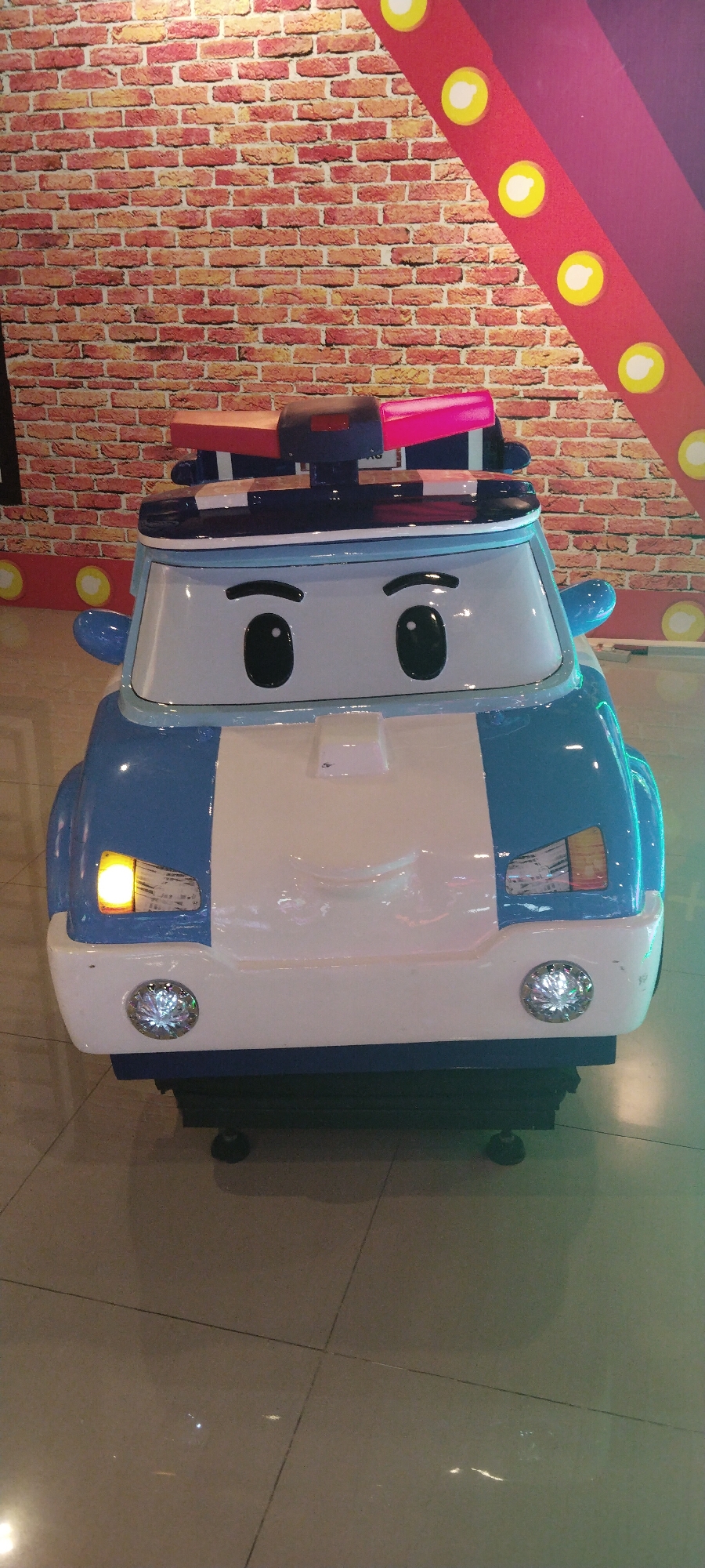 A close-up, eye-level, front-facing shot captures a stylized children's amusement ride resembling a police car. The ride is predominantly white and blue, with large, cartoonish black eyes and eyebrows on the windshield. Red and pink flashing lights are mounted on its roof. The ride sits on a dark base with visible supports. In the background, a red brick wall pattern is visible, along with a section of a decorative, purple and red diagonal banner adorned with yellow circles, likely stage lights or sequins. The ride is positioned on a polished, tiled floor with faint reflections of light. The scene appears to be an indoor entertainment venue, possibly an arcade or amusement park, in Semarang, Indonesia.