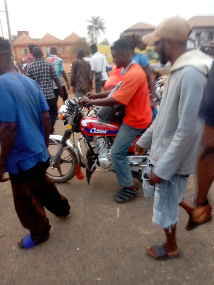 The image depicts an outdoor daytime scene on what appears to be a street or path, bustling with several people and featuring a red motorcycle prominently in the mid-ground. The lighting suggests a bright, possibly overcast day.
In the immediate foreground on the left, a person is seen from the back, wearing a blue short-sleeved t-shirt and dark, possibly brown or black, baggy pants. They are wearing bright blue slide sandals on their feet and are walking towards the right side of the frame.
Central to the image, a person is seated on a red motorcycle, facing away from the camera but with their profile partially visible. This individual is wearing an orange short-sleeved t-shirt, blue jeans, and dark slide sandals with white stripes. Their hands are on the handlebars of the motorcycle, suggesting they are either preparing to ride or are currently stationary. The motorcycle itself is red with a black seat and features chrome elements, including the front forks and engine casing. A yellow circular reflector is visible on the front fork. On the side of the motorcycle's fuel tank, the brand name "Calink" is clearly visible in white and blue lettering.
To the right of the motorcycle, another person stands, facing left towards the person on the motorcycle. This individual is wearing a light grey hooded jacket (hoodie) over a t-shirt, and light blue distressed denim shorts. They are also wearing dark slide sandals. A light brown baseball cap covers their head, and they have a visible beard.
Behind the person on the motorcycle, a partial view of another person in a blue top is visible, possibly a passenger or someone standing close by. Further into the background, several other people are walking or standing. One person in the mid-background wears a plaid shirt, another a white shirt, and others are in various darker attire.
The background features several light-colored buildings with distinct, possibly tiled, roofs, some of which have a rounded or dome-like shape. A tall palm tree is visible in the distance, suggesting a tropical or subtropical setting. The ground beneath the people and motorcycle appears to be asphalt or a hard-packed dirt surface. The overall atmosphere suggests a casual public gathering or daily street activity.