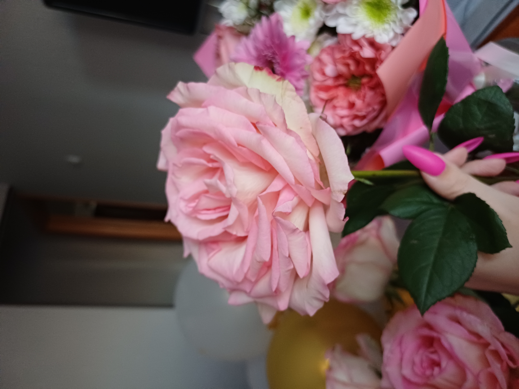 A close-up, eye-level shot captures a bouquet of pink and white flowers. The primary subject is a large, pale pink rose in the foreground, its delicate petals unfurling. To the left of this rose, a hint of a yellow balloon is visible. Behind the rose, other flowers in shades of pink and white, along with green leaves, form a soft background. A hand with bright pink fingernails is holding the stem of the rose. The environment appears to be indoors, with a gray wall and a door visible in the blurred background.