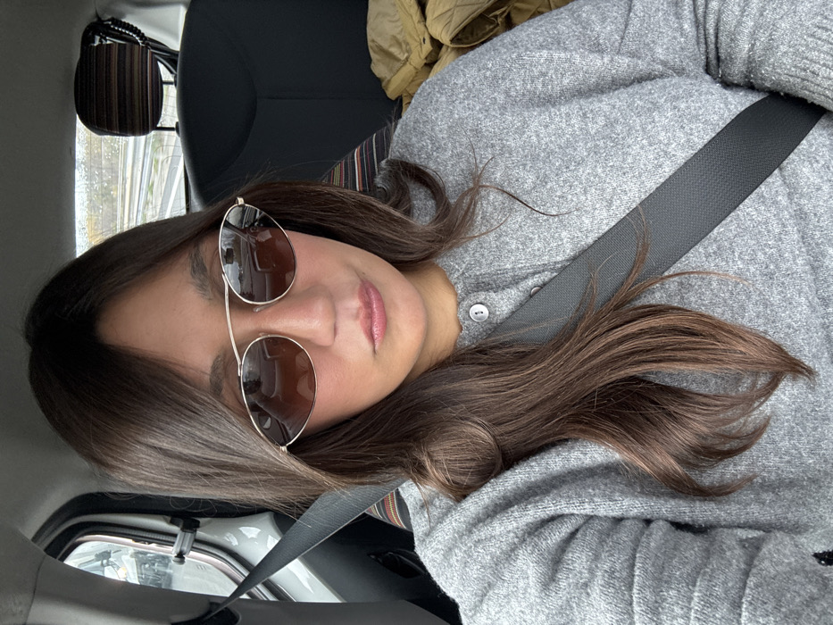 A female subject, located in Vidauban, France, is seated inside a vehicle, likely occupying a front seat. She possesses long, dark brown hair parted centrally, which frames her face. She wears gold-rimmed sunglasses with brown-tinted lenses, obscuring her eyes. Her lips are visible, appearing to be coated with a light pink cosmetic, and are slightly puckered. She is dressed in a light gray, possibly knitted, button-front cardigan or sweater, fastened with light-colored buttons. A black seatbelt is securely buckled across her left shoulder and chest. The interior of the vehicle shows a dark fabric seat and a headrest with a multi-colored striped cover behind her. A beige or tan quilted item is visible on the seat to her right. Through the rear window, a concrete road barrier and vegetation are discernible, indicating a highway or main road setting under daylight conditions.