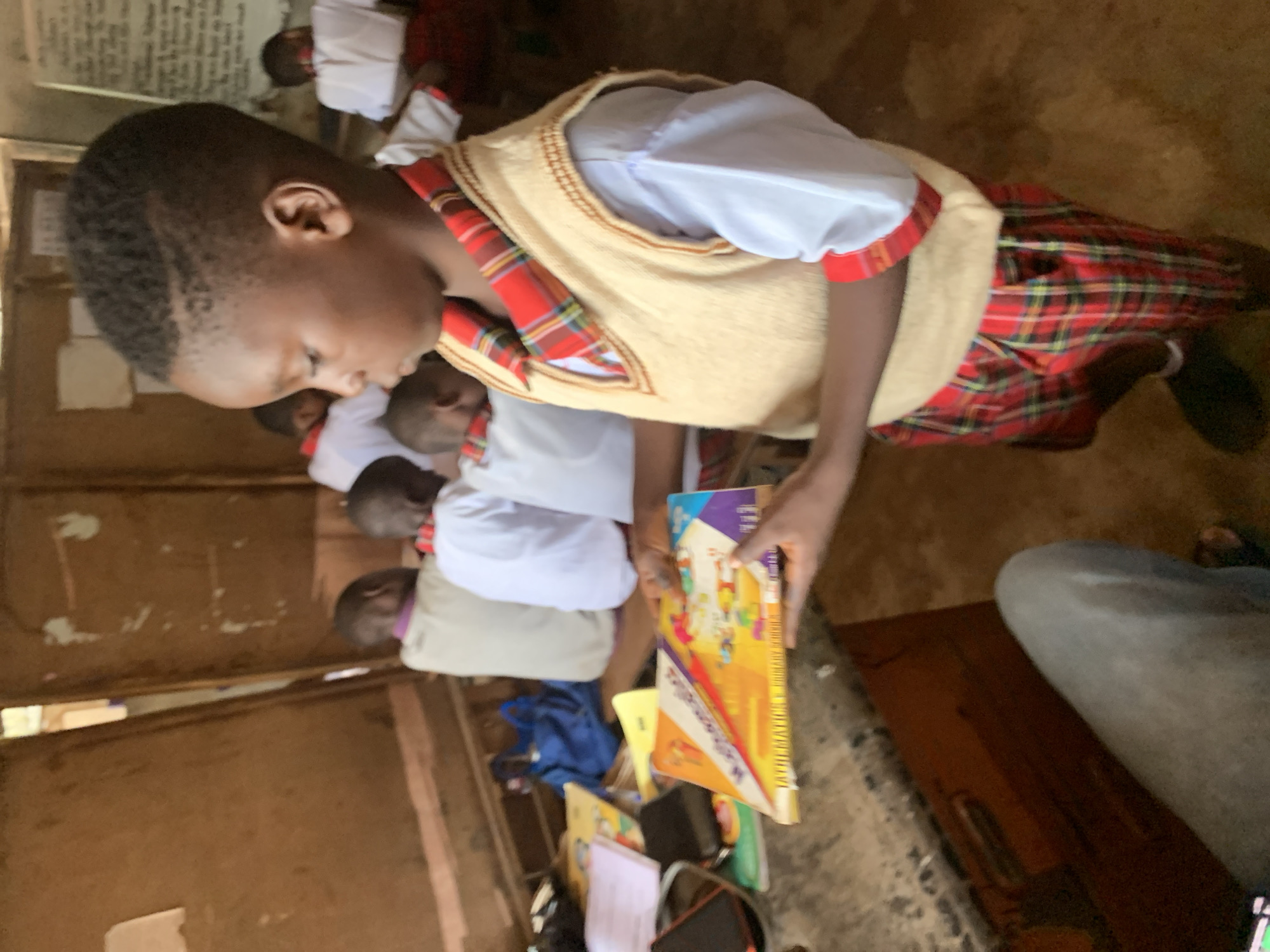 The image shows a classroom scene in Ukwu-Abwa, Nigeria. A young boy, likely a student, is holding a colorful textbook titled "Mathematics." He is wearing a white shirt, a tan vest, and a red plaid skirt or shorts. Several other children, also in school uniforms, are seated in the background. The classroom appears modest with wooden walls and possibly a dirt floor. Other books and items are piled on a surface next to the boy. A portion of a person's leg, clad in jeans, is visible in the lower right corner of the image.