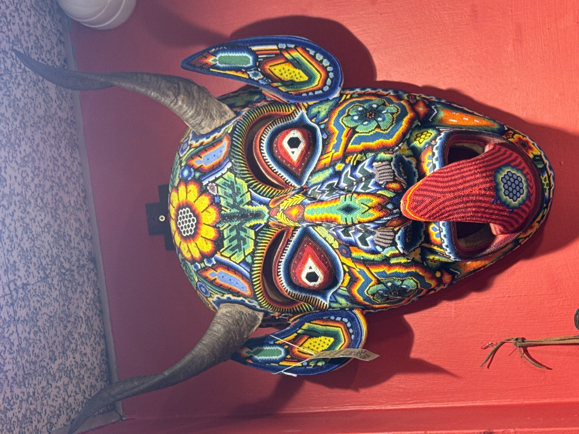 A brightly colored, intricately beaded mask of a devil is affixed to a vibrant red wall. The mask features two curved horns, large expressive eyes outlined in red and white beads, and a long, red, beaded tongue sticking out from its mouth. The mask is adorned with various floral and geometric patterns in a multitude of colors, including yellow, blue, green, and orange. A small, light-colored tag hangs from the left side of the mask. The wall appears to be a smooth, painted surface. Above the mask, a textured ceiling is visible. The overall scene depicts a piece of decorative art, likely a cultural artifact, displayed indoors. The location is identified as London, United Kingdom.