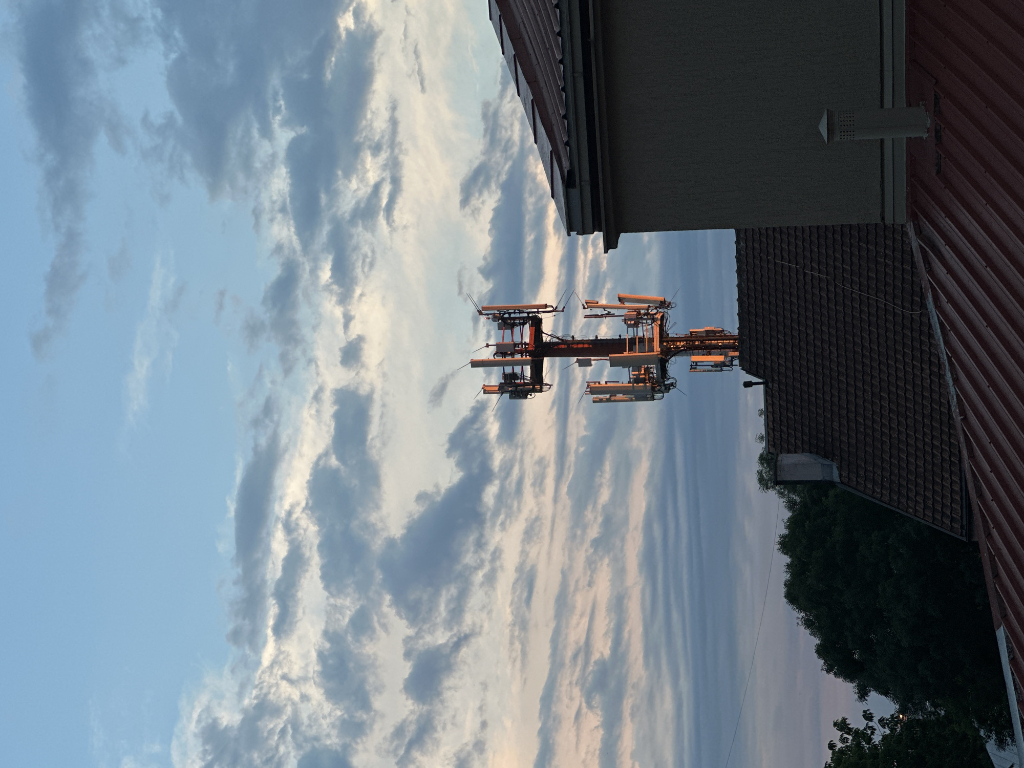 In Vracar Urban Municipality, Serbia, a cell tower stands prominently against a sky filled with wispy clouds painted in hues of blue, white, and pale pink. The tower, a lattice structure, is equipped with multiple antennas, some reflecting the warm glow of the setting sun. In the foreground, the tiled roofs of several buildings, one with a weathered, dark brown finish and another with a reddish-brown metal sheeting, partially obscure the view of the tower and the lower section of a neutral-colored building. A dark green canopy of trees is visible to the left, and a chimney cap adorns a roof on the right. The scene is devoid of human presence.